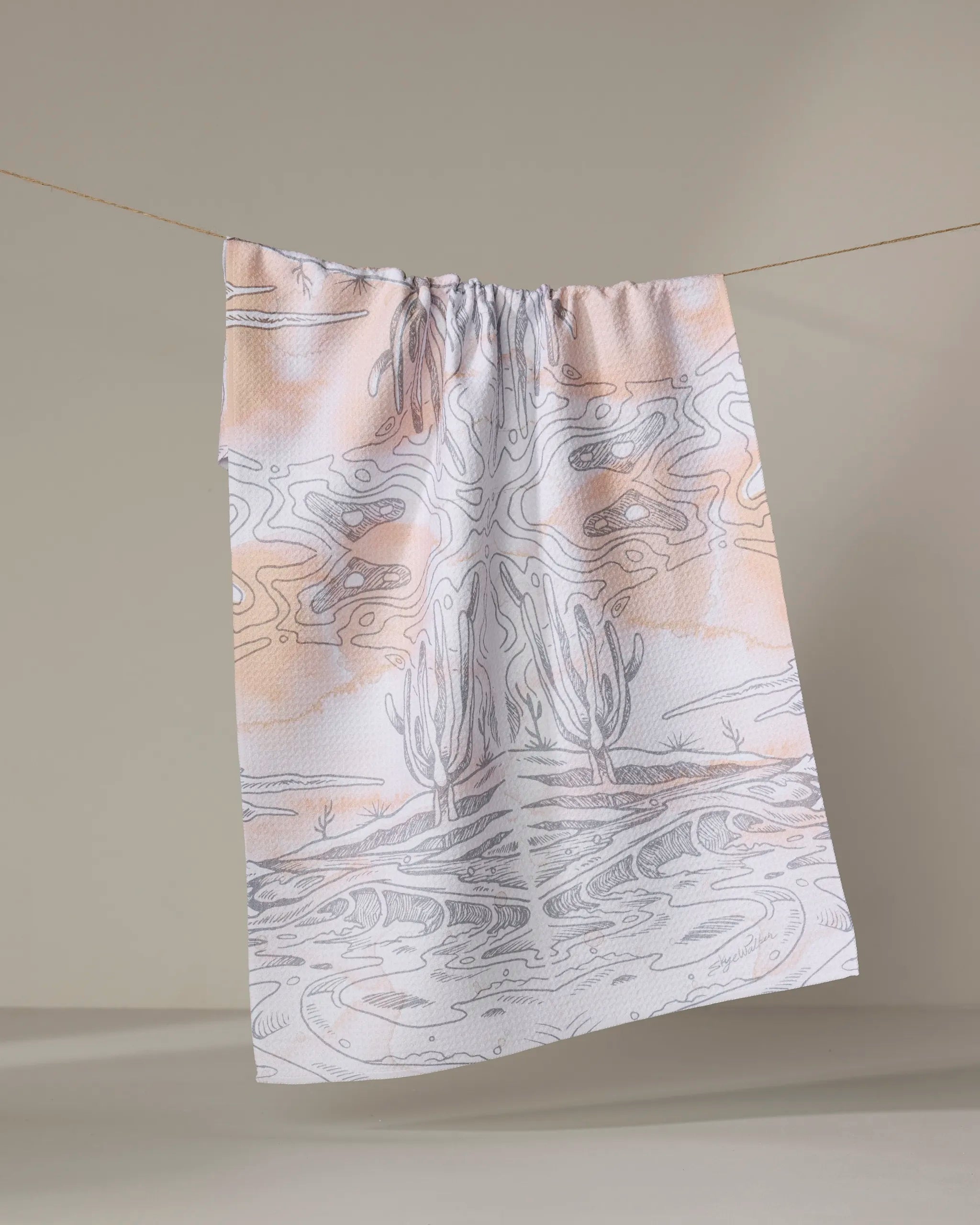 The Skye Walker "Waves" tea towel, crafted from recycled materials, displays a soft pastel desert landscape with cacti, abstract lines, and subtle orange and gray tones, hanging on a string against a neutral background.