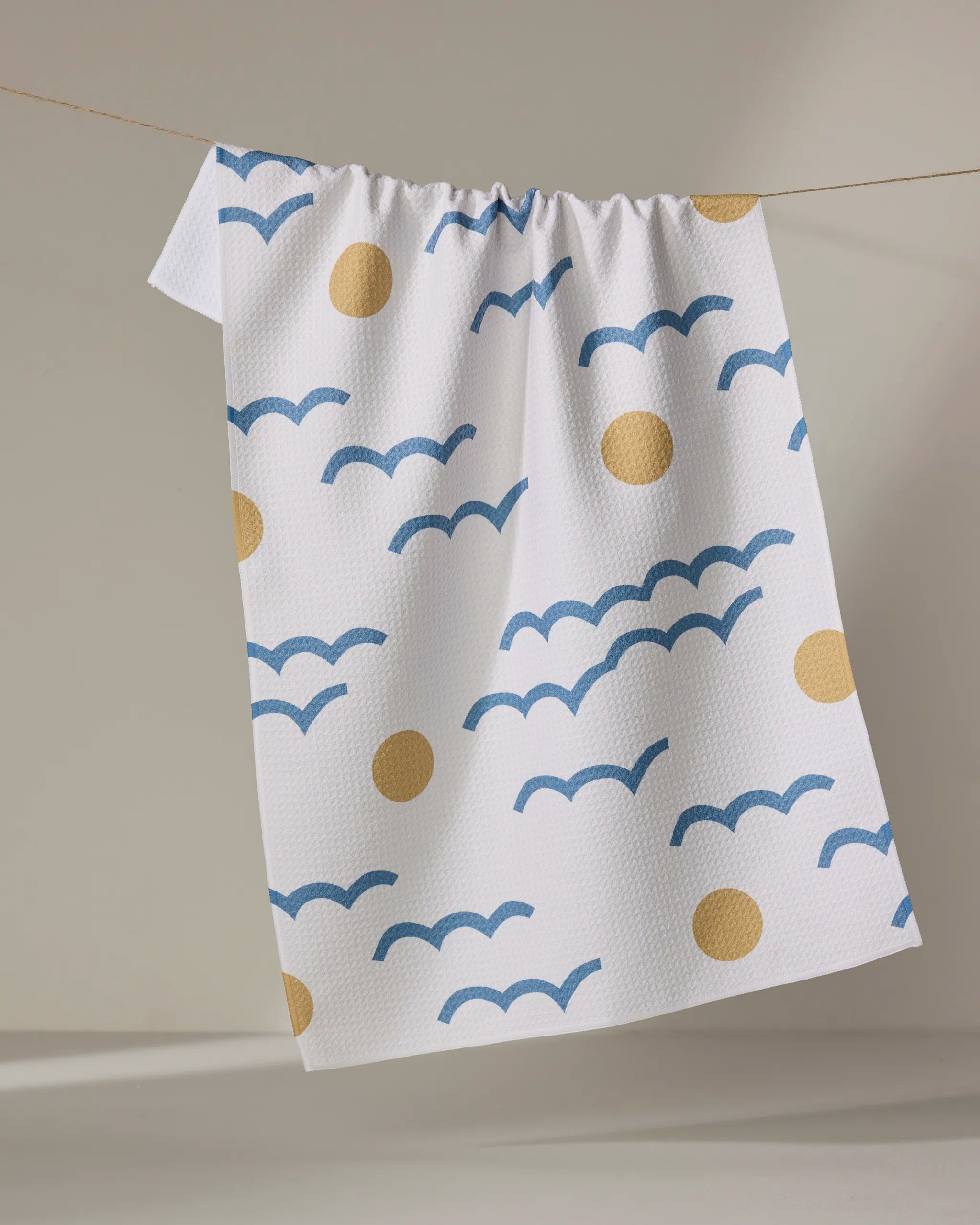 The "Wavy Day" kitchen towel by Elizabeth Edwards is an eco-friendly white towel featuring blue wavy lines and yellow circles, shown hanging on a clothesline against a neutral background.