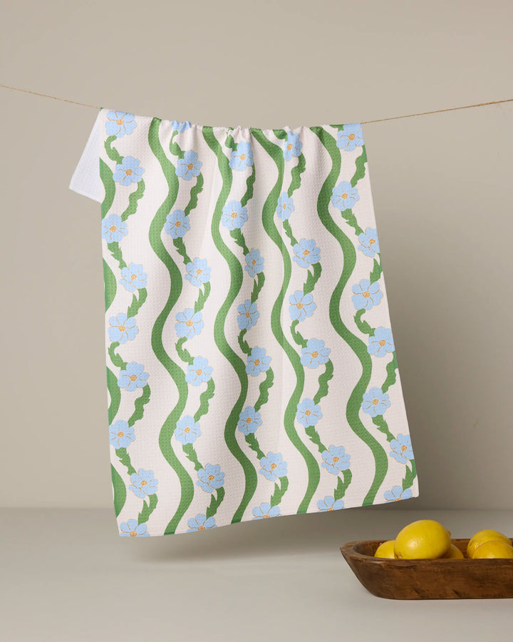 The "Wavy Flowers" tea towel by Geometry, featuring green wavy lines and blue flowers on white, hangs on a string. Three yellow lemons sit in a wooden bowl in front, with a neutral background.