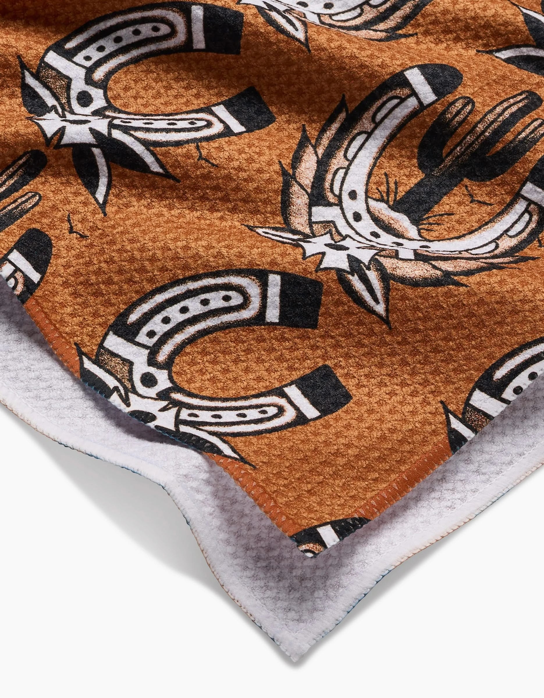 A close-up of Cole Strems Western Horseshoes towel highlights its ultra-absorbent Geoweave fabric in brown, featuring black and white horseshoes and cacti patterns with a white bottom edge.