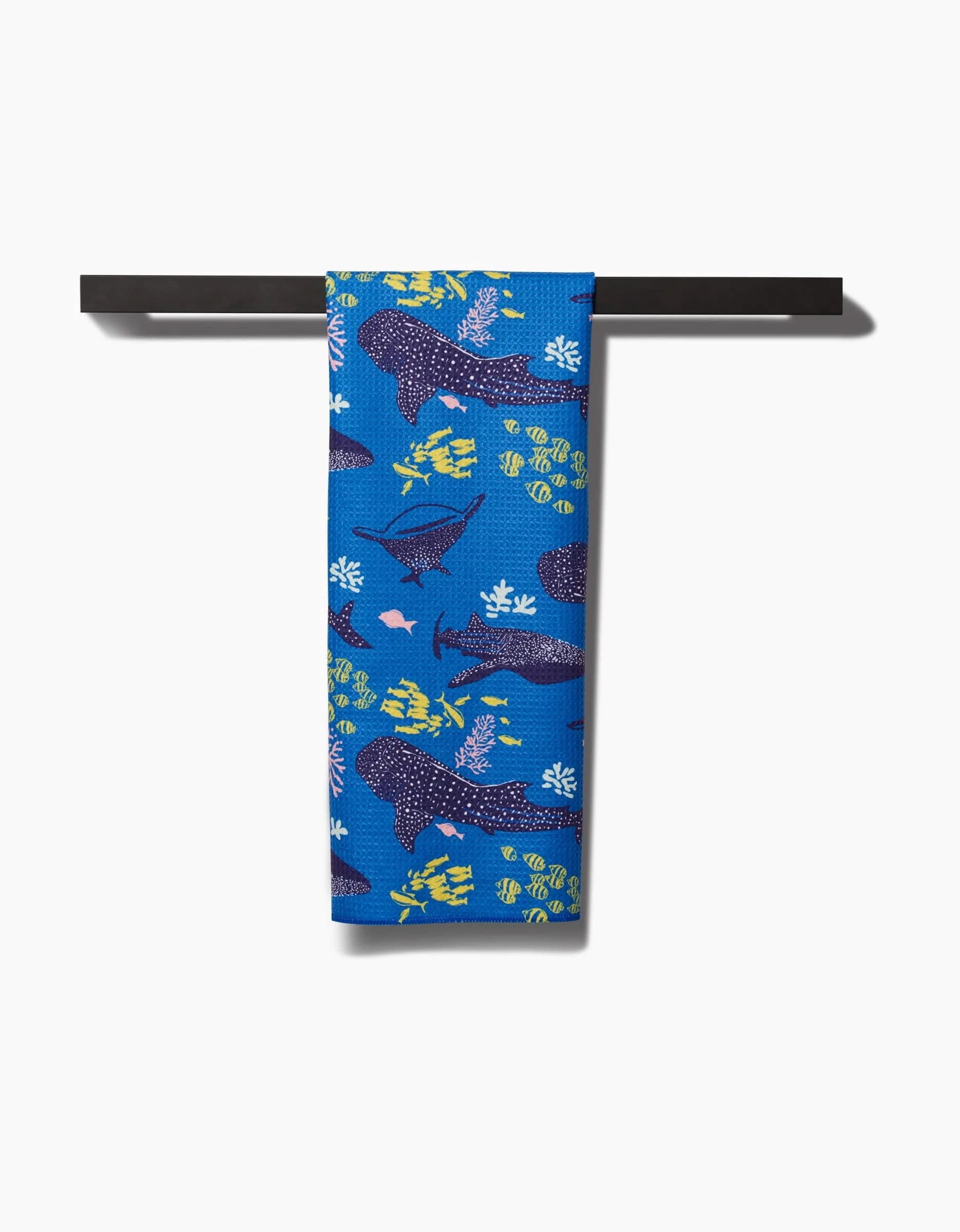 The Elisabeth Wing Whale Shark tea towel hangs on a black bar, displaying whale sharks, tropical fish, and coral in yellow, green, and navy—made from Geoweave™ fabric with post-consumer recycled materials.