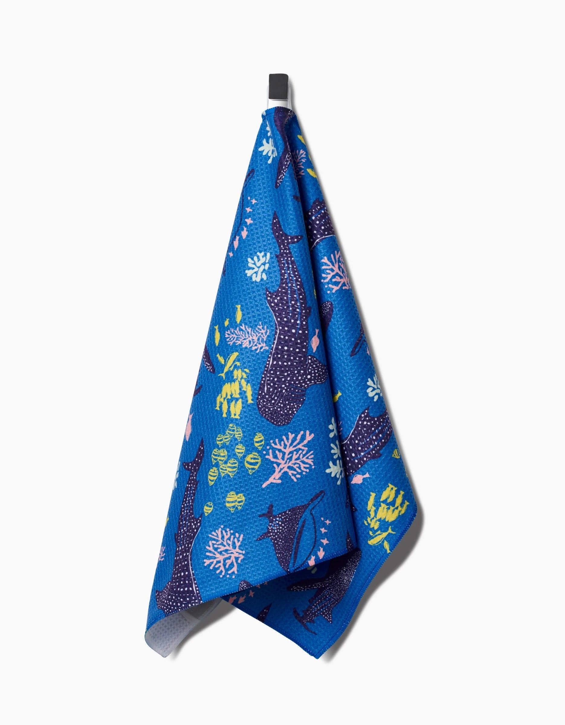 The Whale Shark tea towel by Elisabeth Wing hangs from a hook, showcasing lively ocean-themed patterns with whale sharks, corals, and tropical fish in purple, yellow, pink, and green—crafted from post-consumer recycled materials.