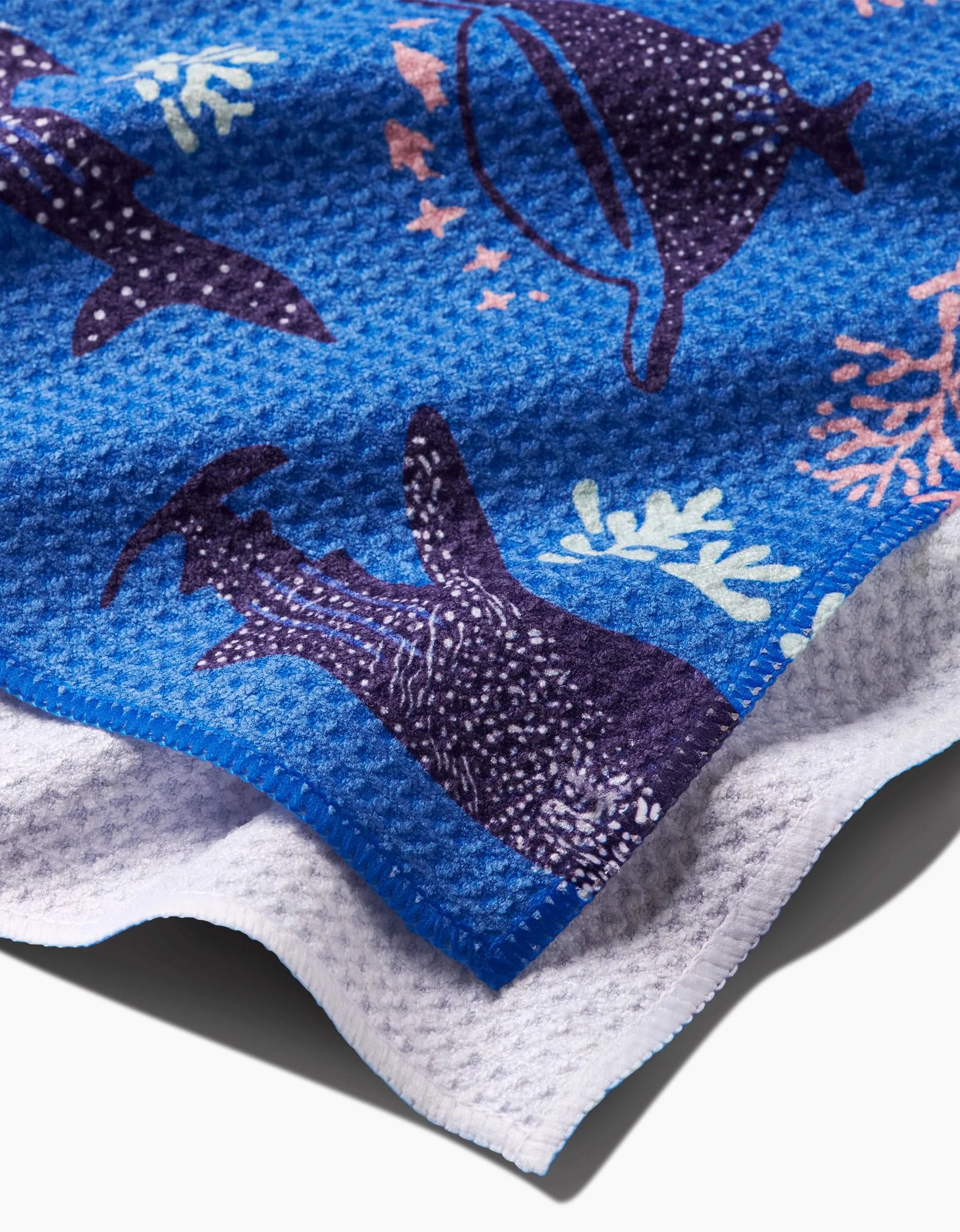 A close-up of the Elisabeth Wing Whale Shark tea towel shows blue textures with whale sharks, corals, and sea stars in dark blue and pastel shades. Made from post-consumer recycled materials, the edge reveals a white underside.