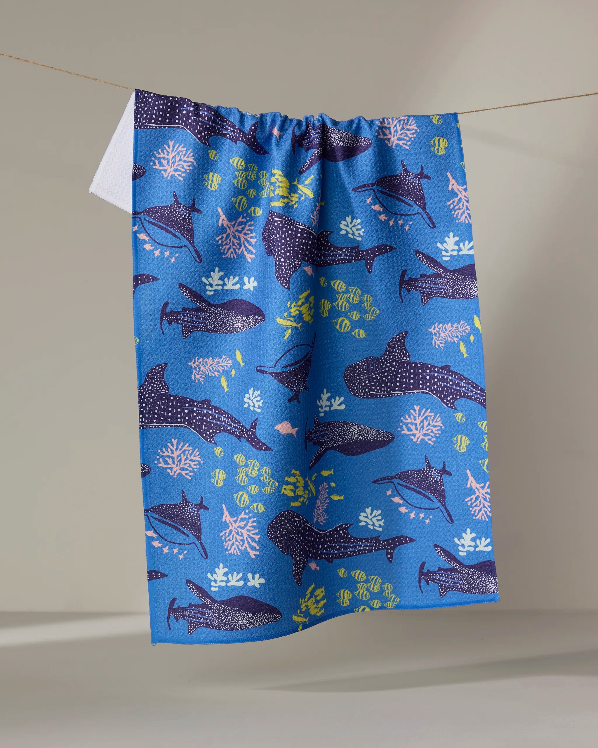 A blue absorbent dish towel by Elisabeth Wing, named Whale Shark, features colorful illustrations of sharks, fish, and coral reefs and hangs from a string against a plain background.