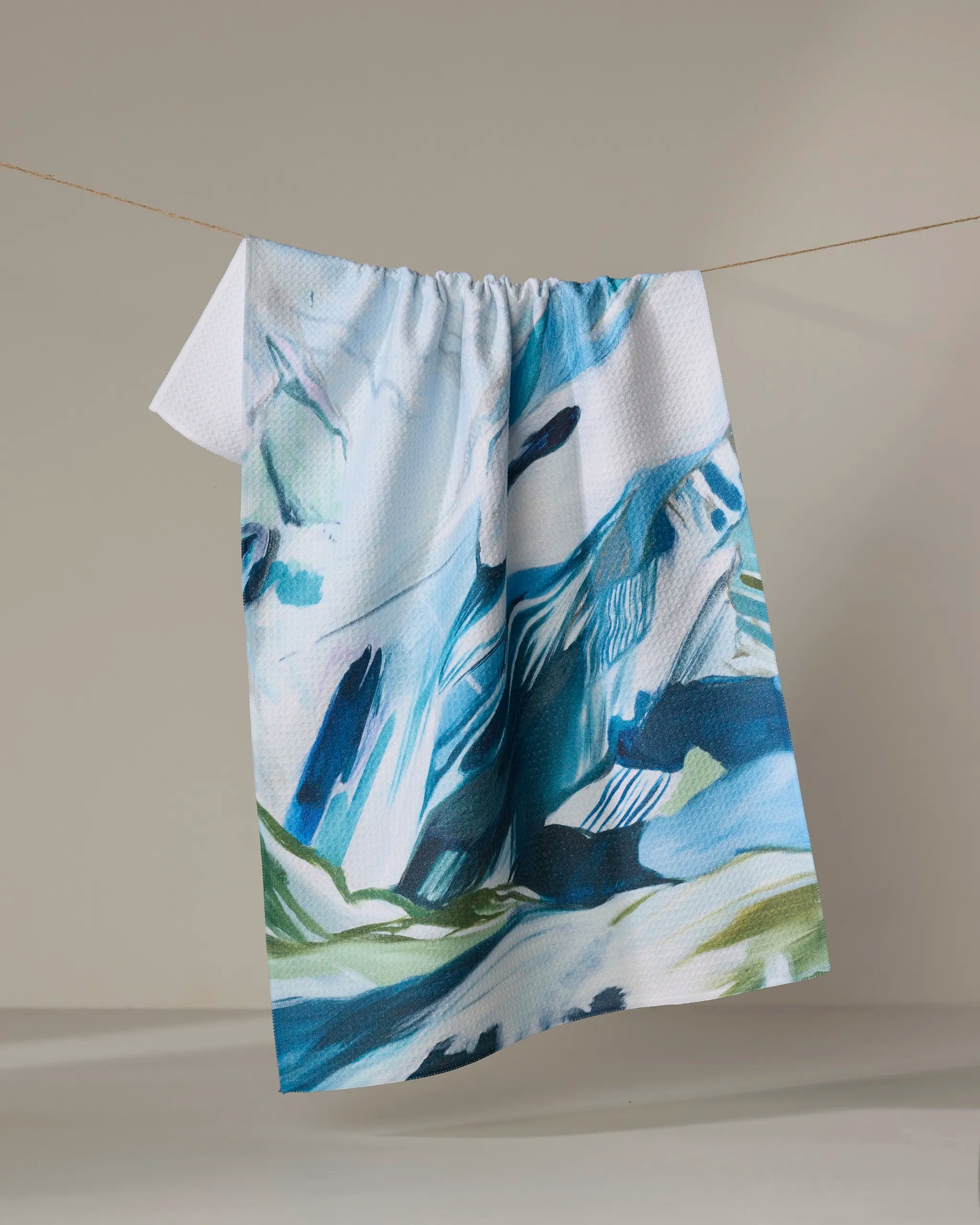 The "Where He Laid To Go" kitchen towel by Kelly Peters features an abstract blue, green, and white design, hangs on a clothesline against a neutral background, and is crafted from recycled materials.