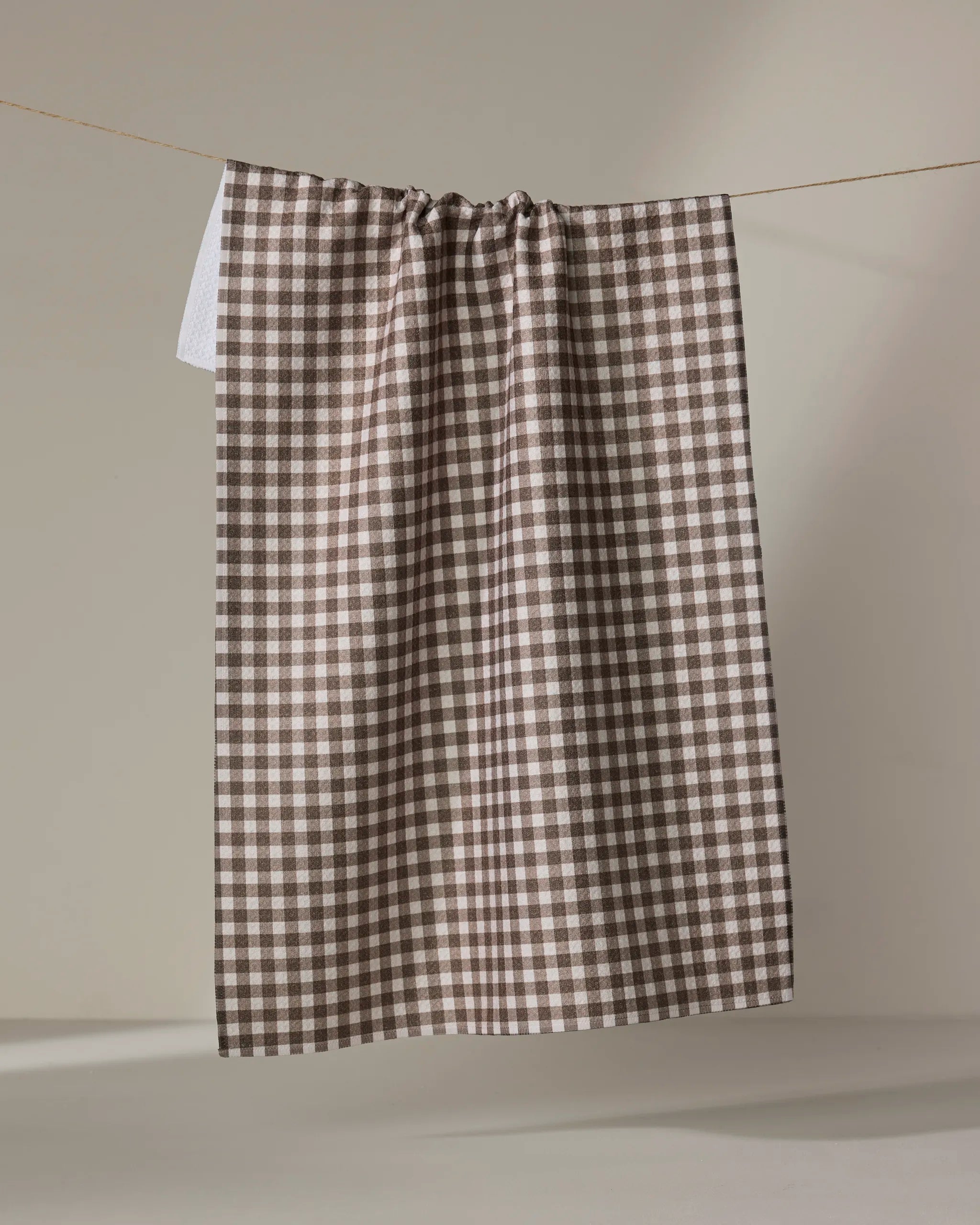 The Whidbey Check tea towel by Jenny Keller, featuring a brown and white checkered design, hangs neatly from a string against a plain neutral background.