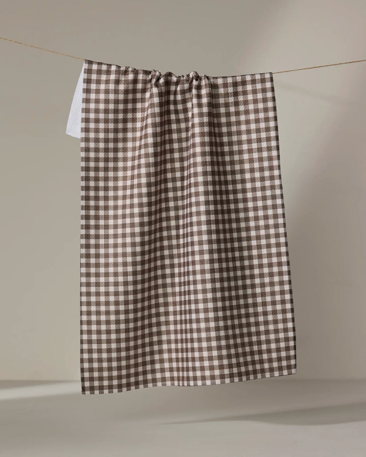 The Whidbey Check tea towel by Jenny Keller, featuring a brown and white checkered design, hangs neatly from a string against a plain neutral background.