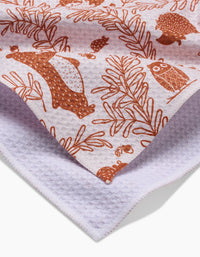 A close-up of the Sara Mari Whimsical Woodland ultra-absorbent bar towel, featuring brown bear, owl, hedgehog, and leaf illustrations on white textured fabric. The folded towel displays its patterned front and plain back.
