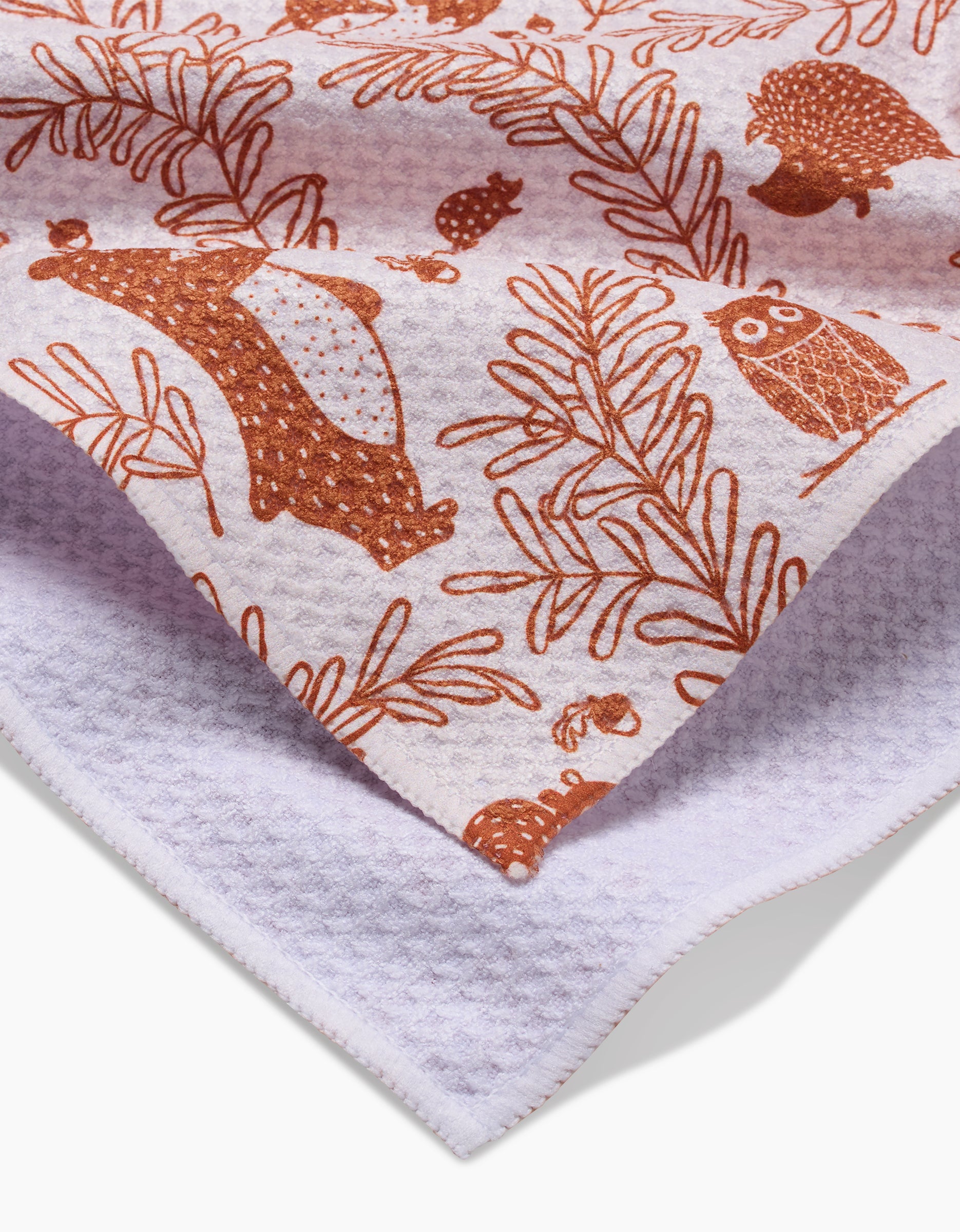 A close-up of the Sara Mari Whimsical Woodland ultra-absorbent bar towel, featuring brown bear, owl, hedgehog, and leaf illustrations on white textured fabric. The folded towel displays its patterned front and plain back.