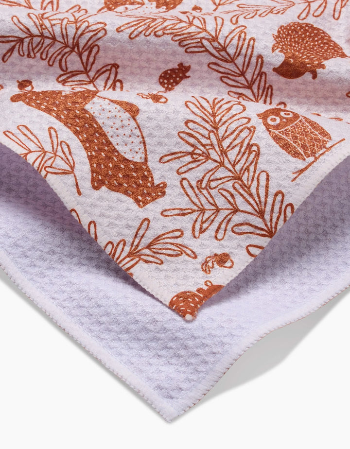 A close-up of the Sara Mari Whimsical Woodland ultra-absorbent bar towel, featuring brown bear, owl, hedgehog, and leaf illustrations on white textured fabric. The folded towel displays its patterned front and plain back.