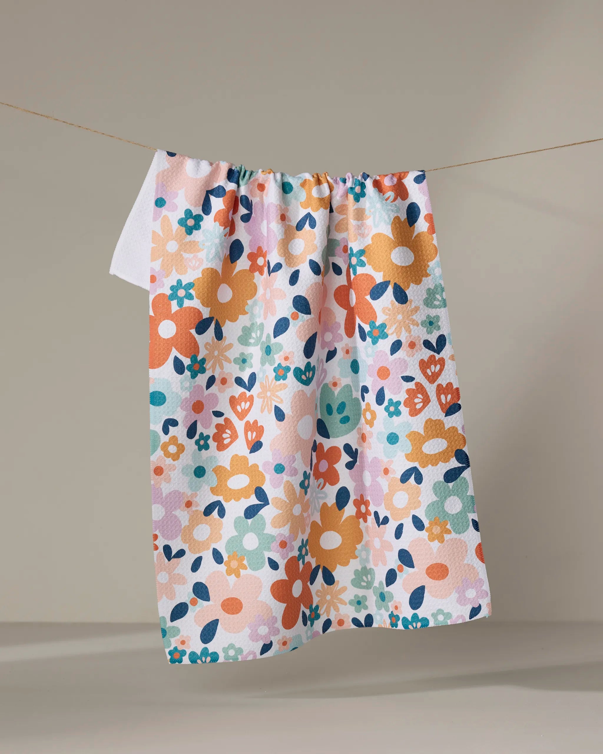 The Sara Mari Whimsy Floral tea towel, made from quick-drying recycled materials, features a playful orange, blue, pink, and green floral design and hangs on a string against a neutral background.