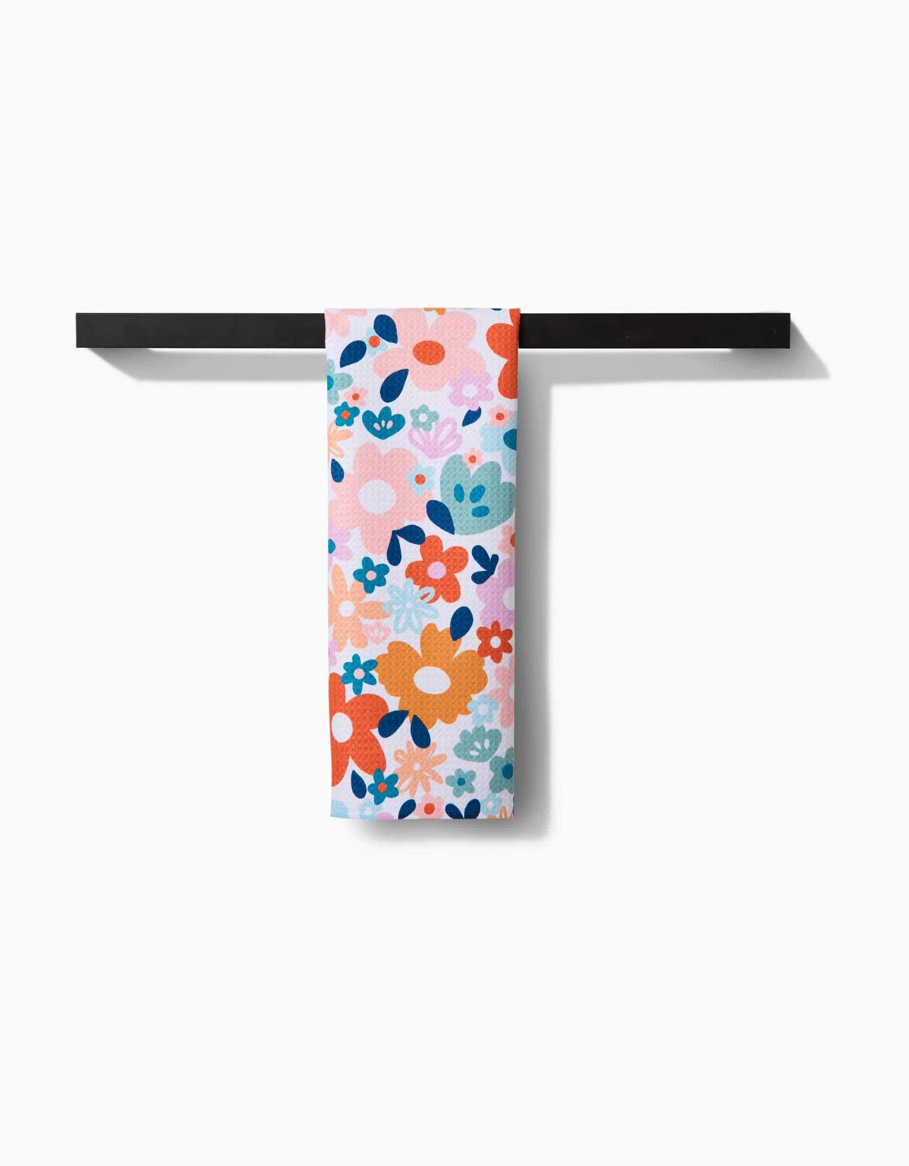 The Sara Mari Whimsy Floral Geometry Tea Towel, featuring a lively orange, pink, blue, and teal floral pattern, hangs neatly on a horizontal black bar with a white background.