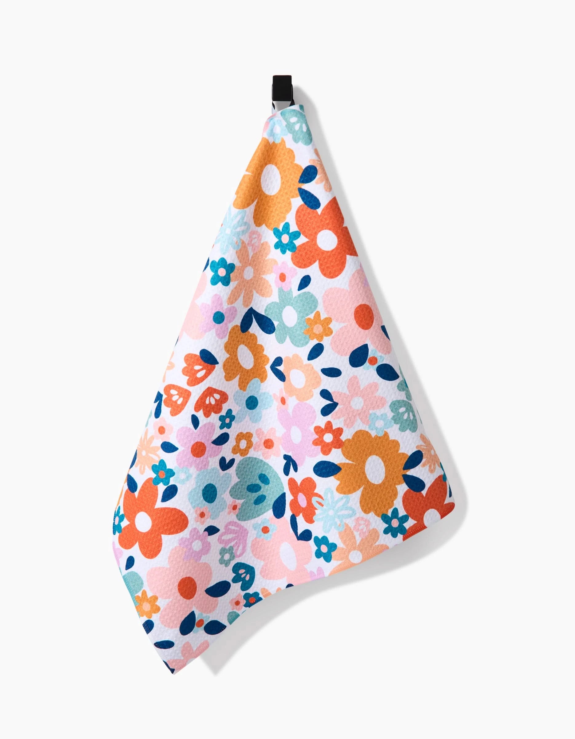 The Sara Mari Whimsy Floral Geometry Tea Towel, featuring a vibrant orange, pink, blue, and green floral pattern, hangs from a black loop against a plain white background. Made with Geoweave fabric for superior absorbency.