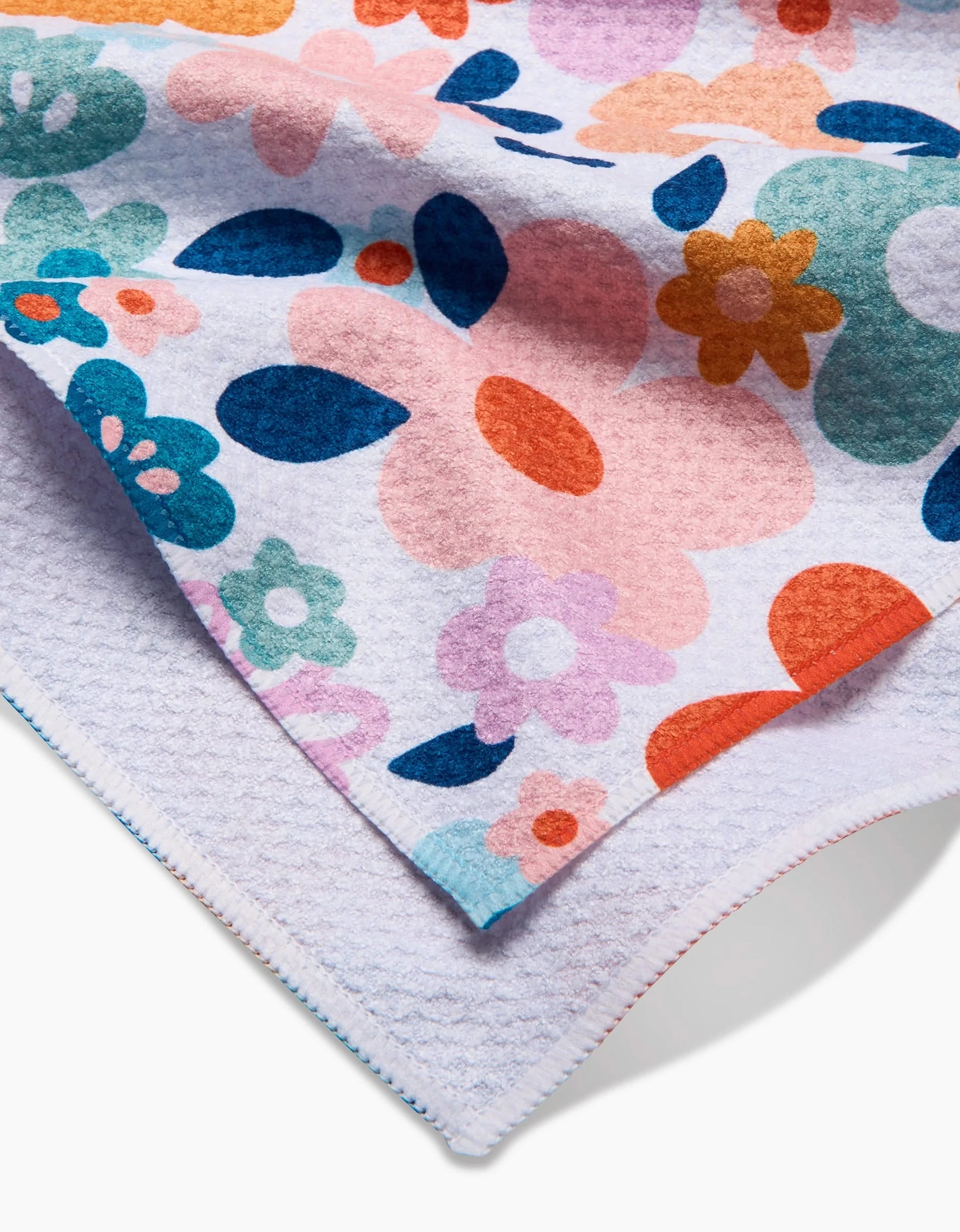 A close-up of the Whimsy Floral tea towel by Sara Mari, crafted from recycled materials with a bold floral pattern in pink, orange, blue, green, and purple on one side and a plain white textured surface on the other.