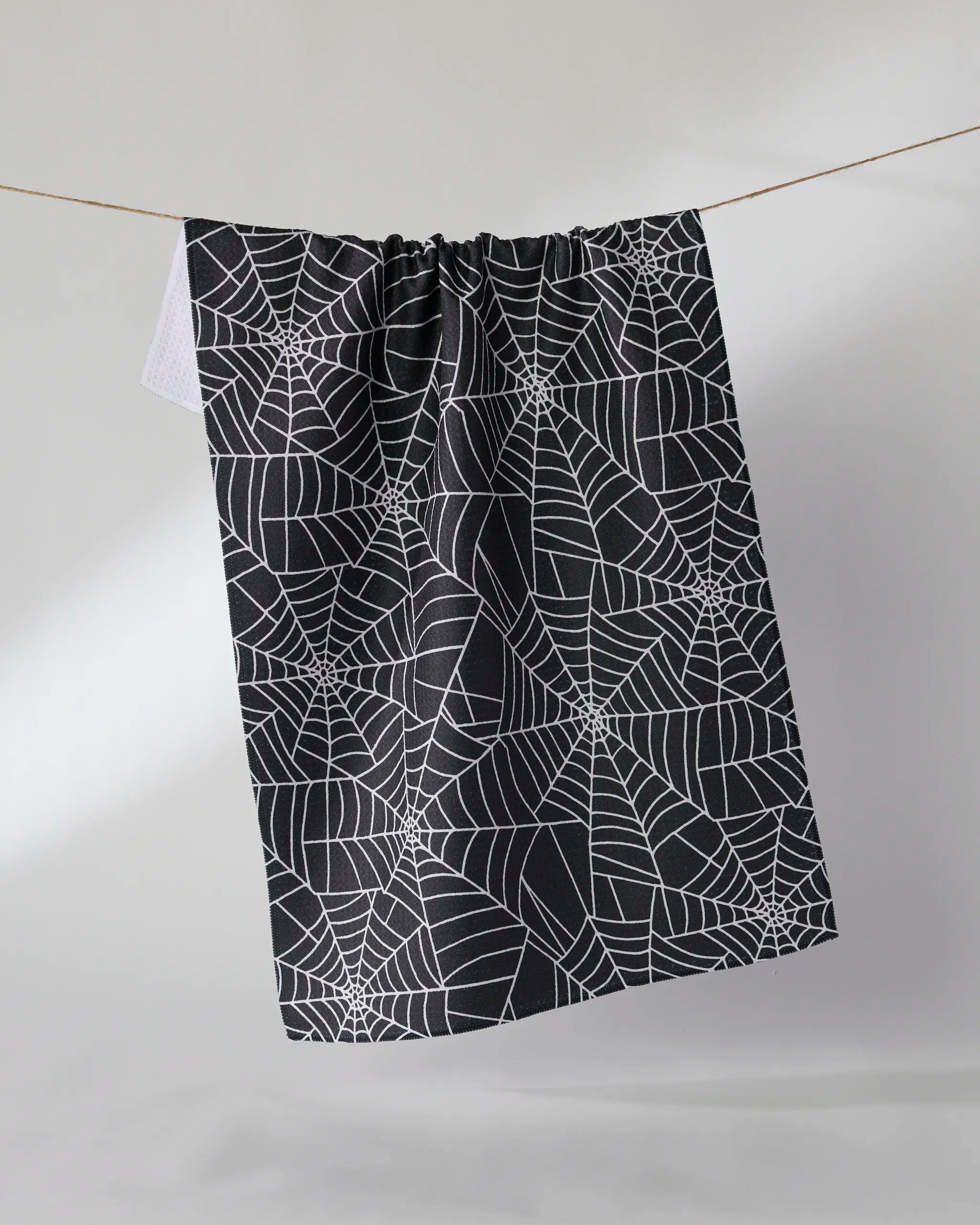 The Geometry Whisper Web dish towel, featuring a black absorbent fabric with a white spiderweb pattern, hangs on a thin string against a plain light background.