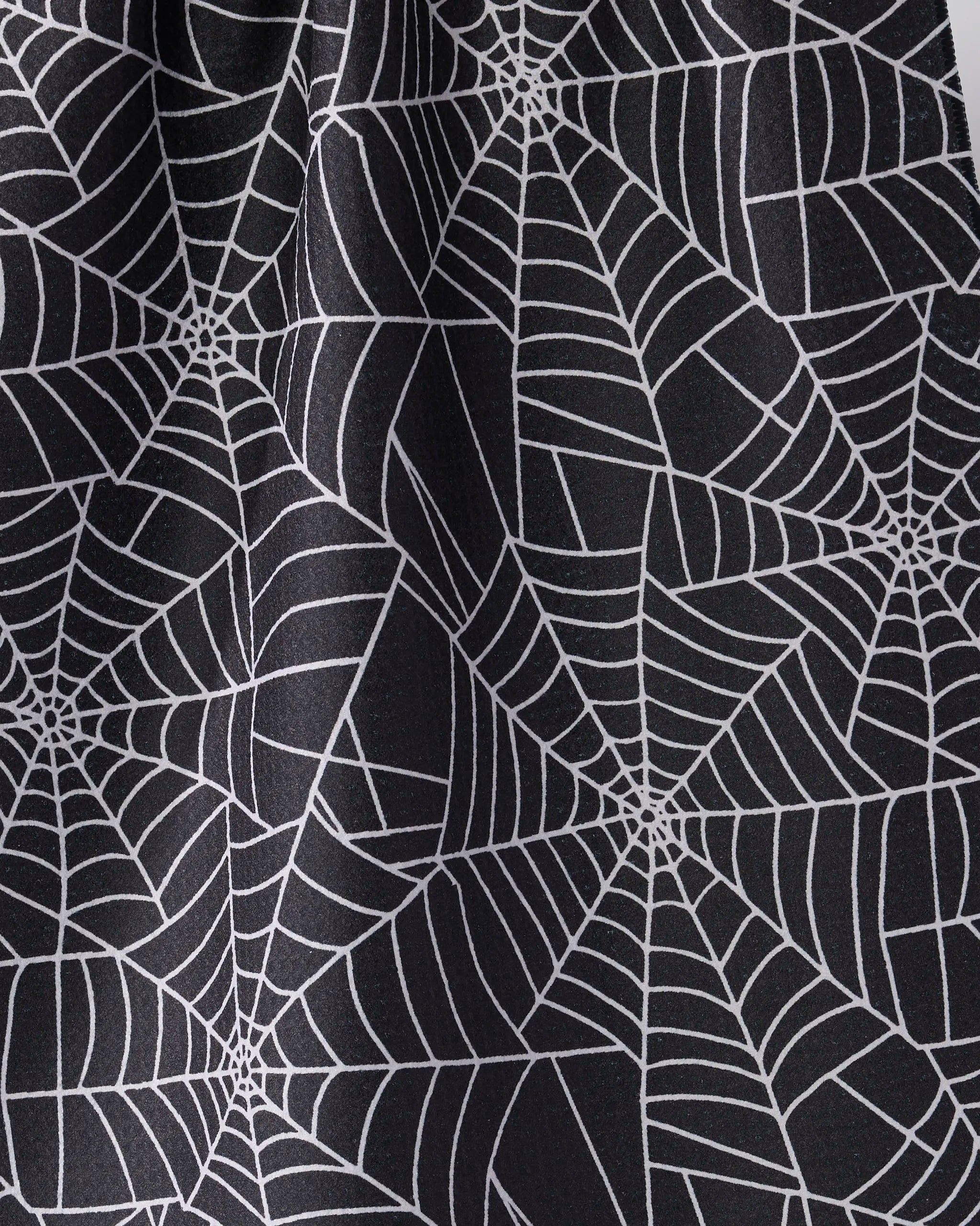 A close-up of Geometry's Whisper Web kitchen towel shows black fabric with intersecting white spider web patterns. This eco-friendly towel features a unique design and a softly wrinkled texture.