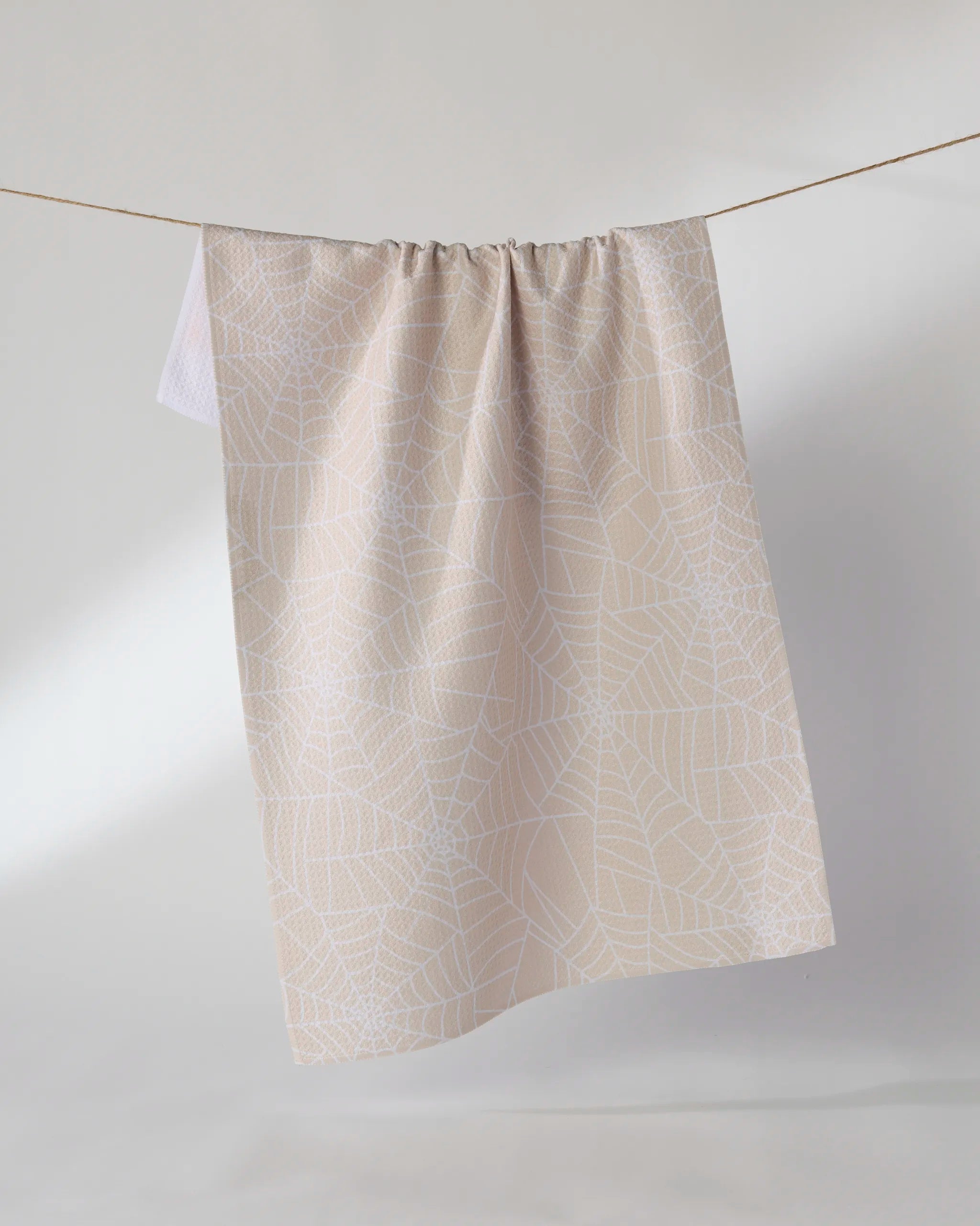 The Geometry Whisper Web kitchen towel, in beige with a subtle white spiderweb pattern, hangs neatly on a clothesline against a plain, light background.