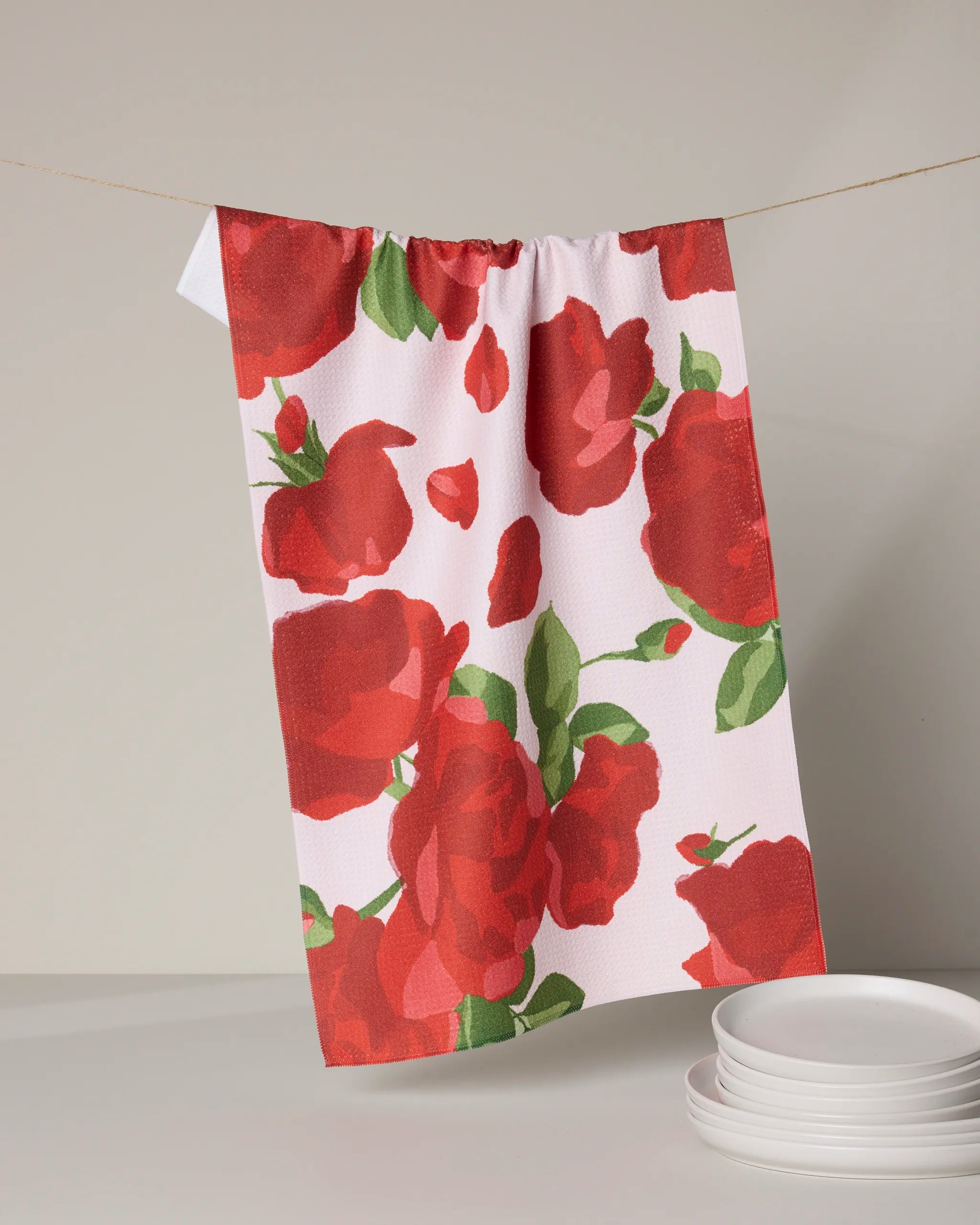 The Geometry Whispering Roses white tea towel, featuring a bold red rose and green leaf design, hangs on a line. Quick-drying and ideal for daily use, it’s shown above a stack of white plates.