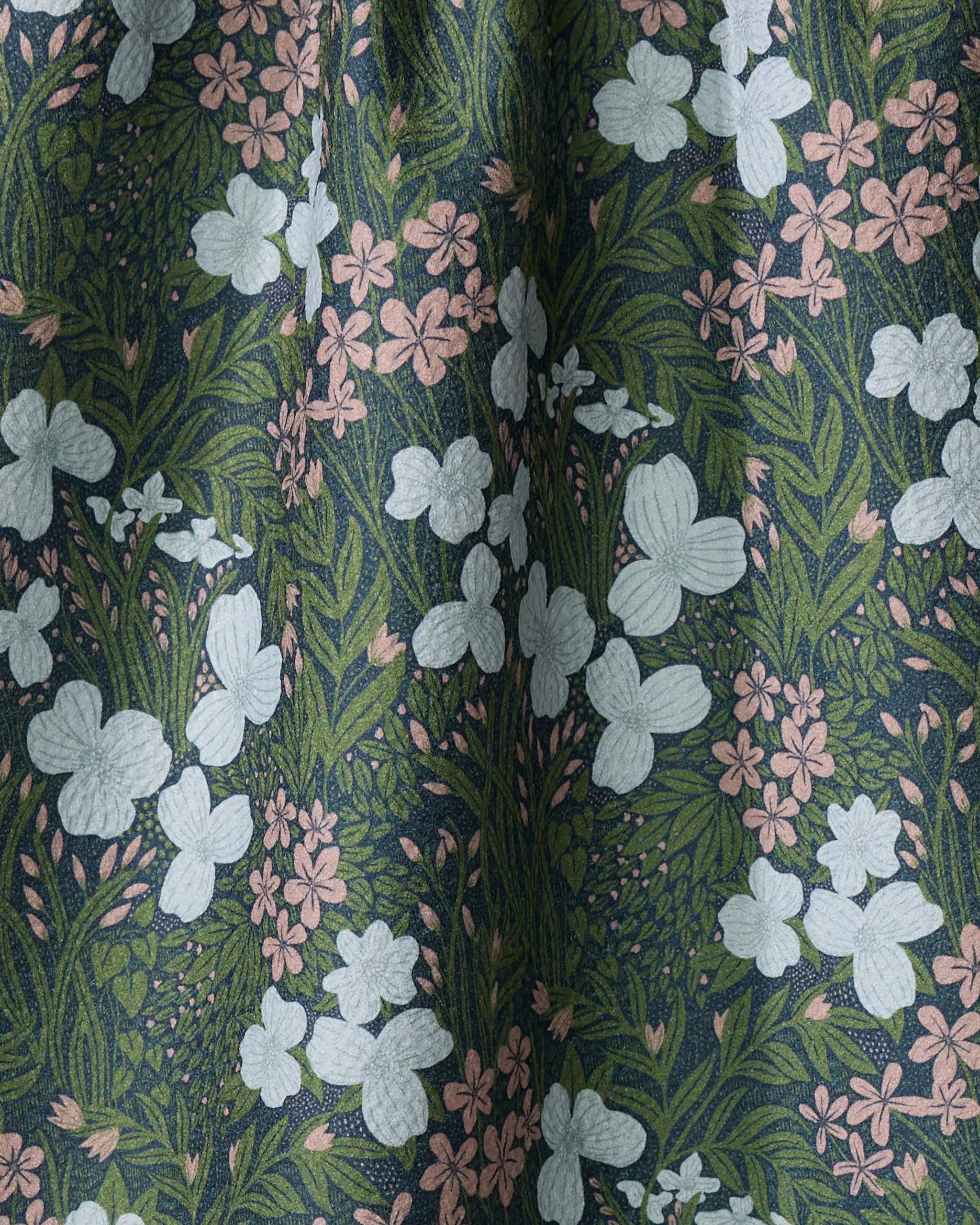 A close-up of the White And Green Meadow kitchen towel by Katie O'Shea shows a floral print with white and pale pink flowers and green leaves on dark fabric, slightly gathered for charm. Made from recycled materials.