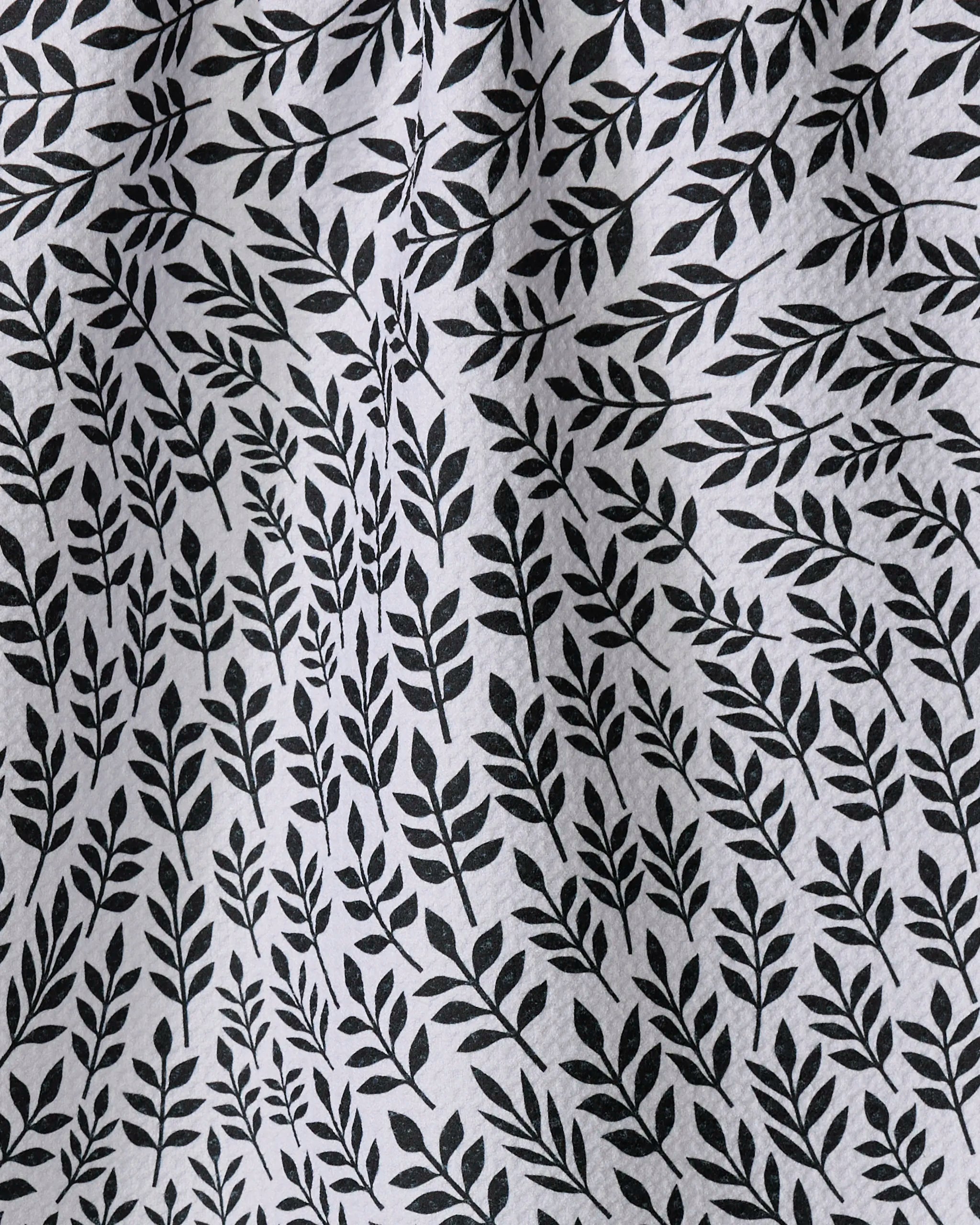 A close-up of the Rachel Parker White Leaf Swirl tea towel shows its black leafy branch pattern. The slightly wrinkled fabric creates subtle shadows and texture over the repeating botanical design.