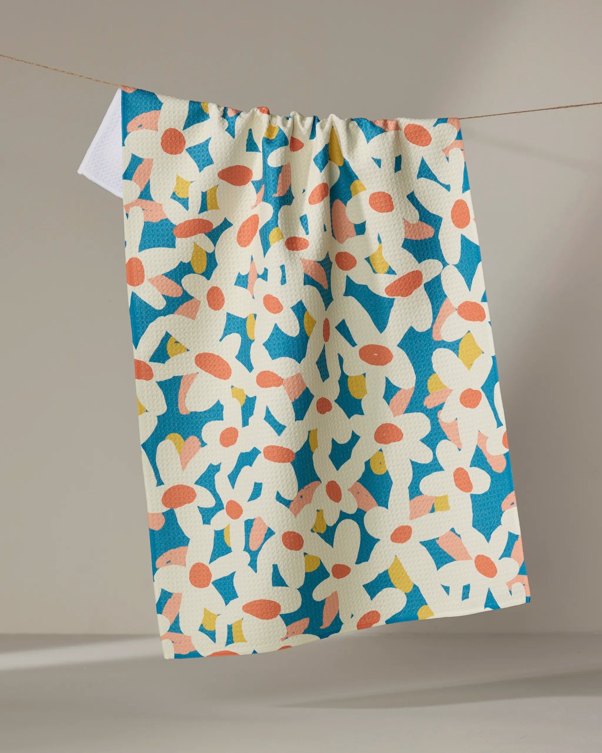 The Whoopsie Daisy tea towel by Kelsey Weigl, featuring a playful orange, yellow, white, and blue floral pattern, drapes over a line against a neutral background.