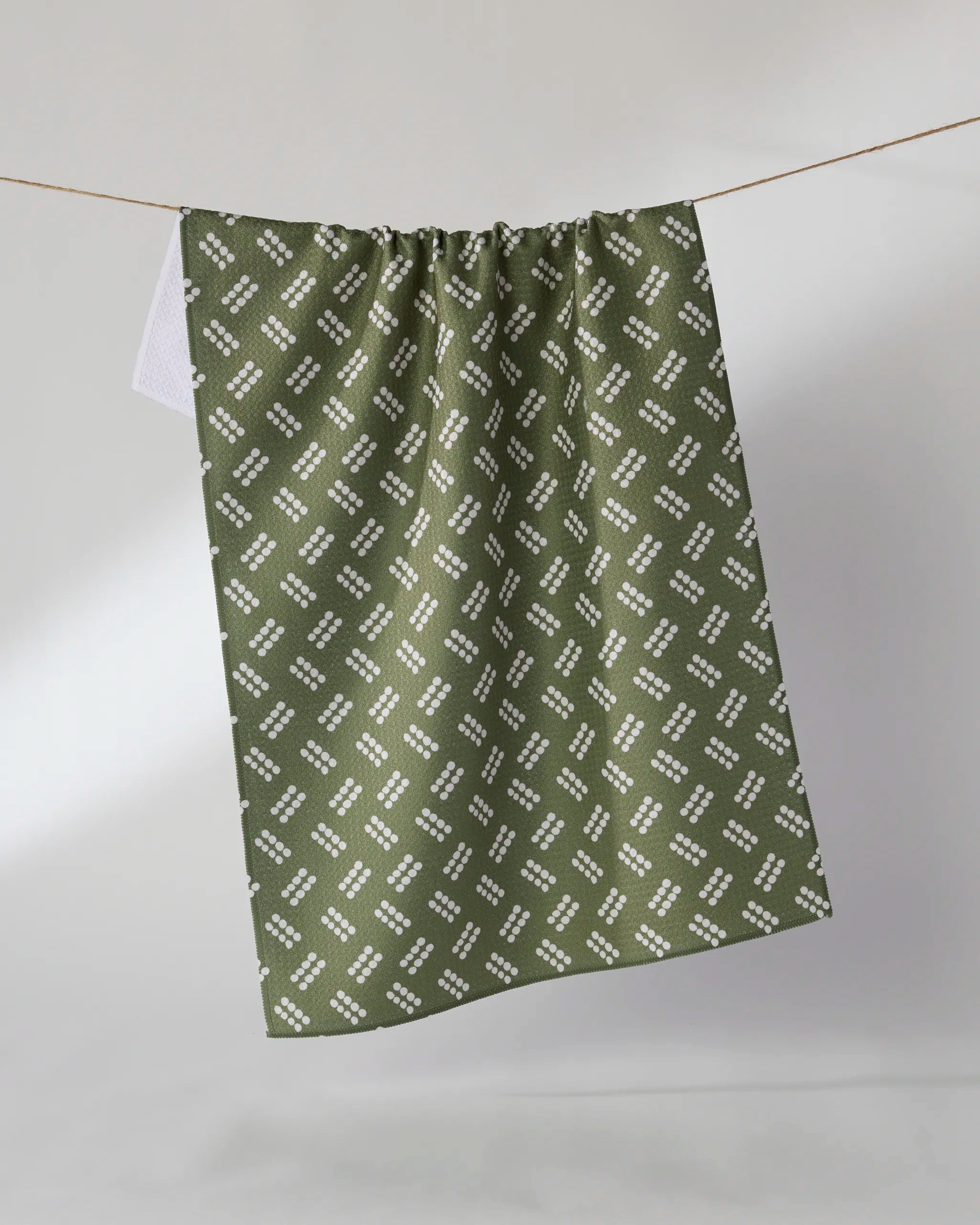 The Geometry Wick tea towel, featuring a white geometric diamond pattern on green, hangs on a line against a light gray background.