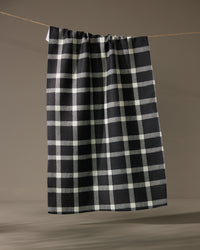 The Geometry Wicklow Black kitchen towel, featuring a black and white checkered pattern, hangs on a clothesline against a simple, neutral background.