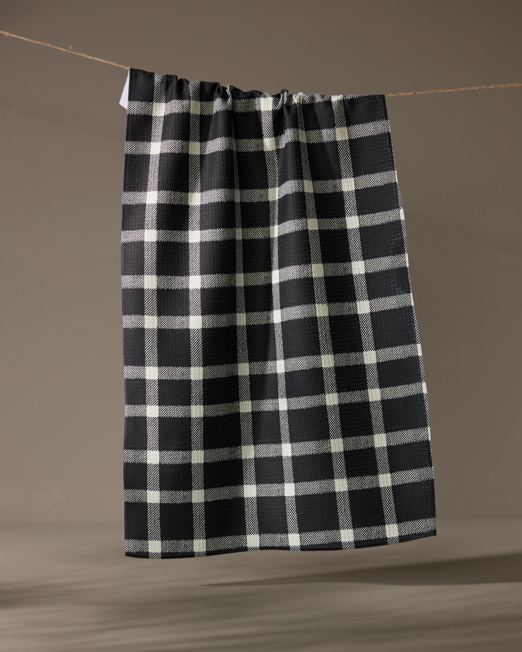 The Geometry Wicklow Black kitchen towel, featuring a black and white checkered pattern, hangs on a clothesline against a simple, neutral background.