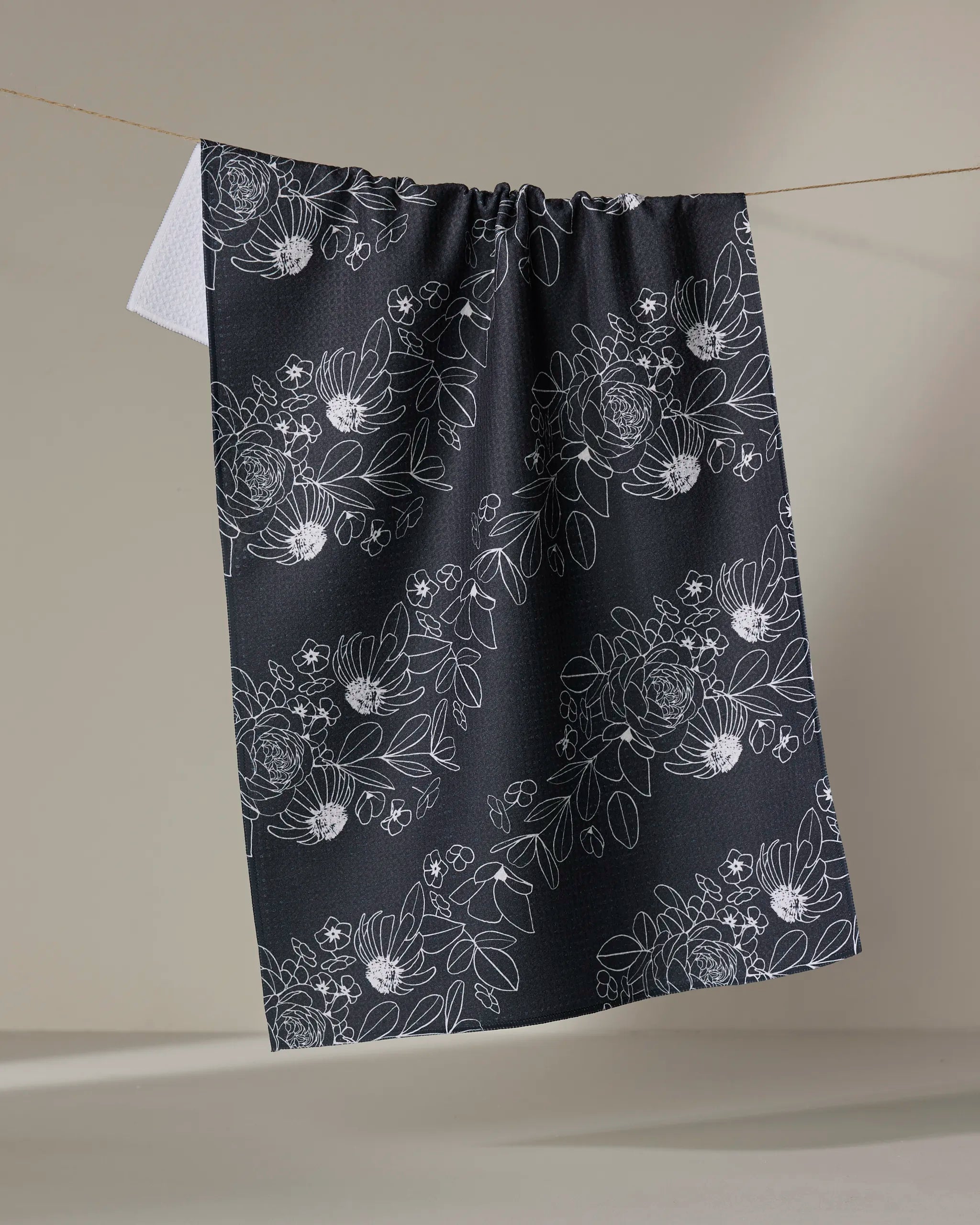 The "Wild and Wonderful" tea towel by Rosa Kichline features a white floral pattern on dark fabric, hung against a neutral background to highlight its design—an elegant addition to any kitchen.