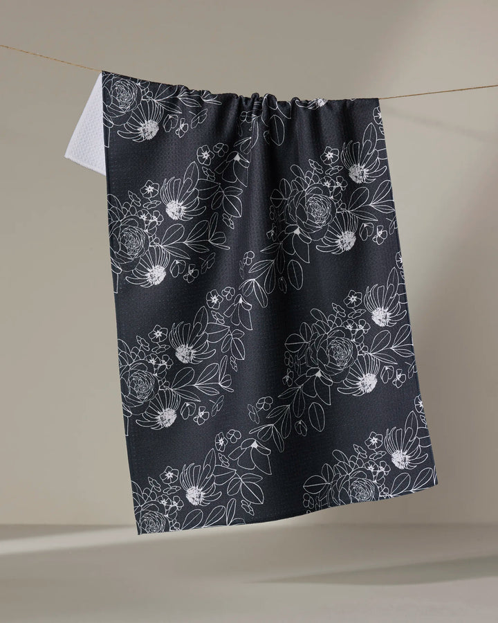 The "Wild and Wonderful" tea towel by Rosa Kichline features a white floral pattern on dark fabric, hung against a neutral background to highlight its design—an elegant addition to any kitchen.