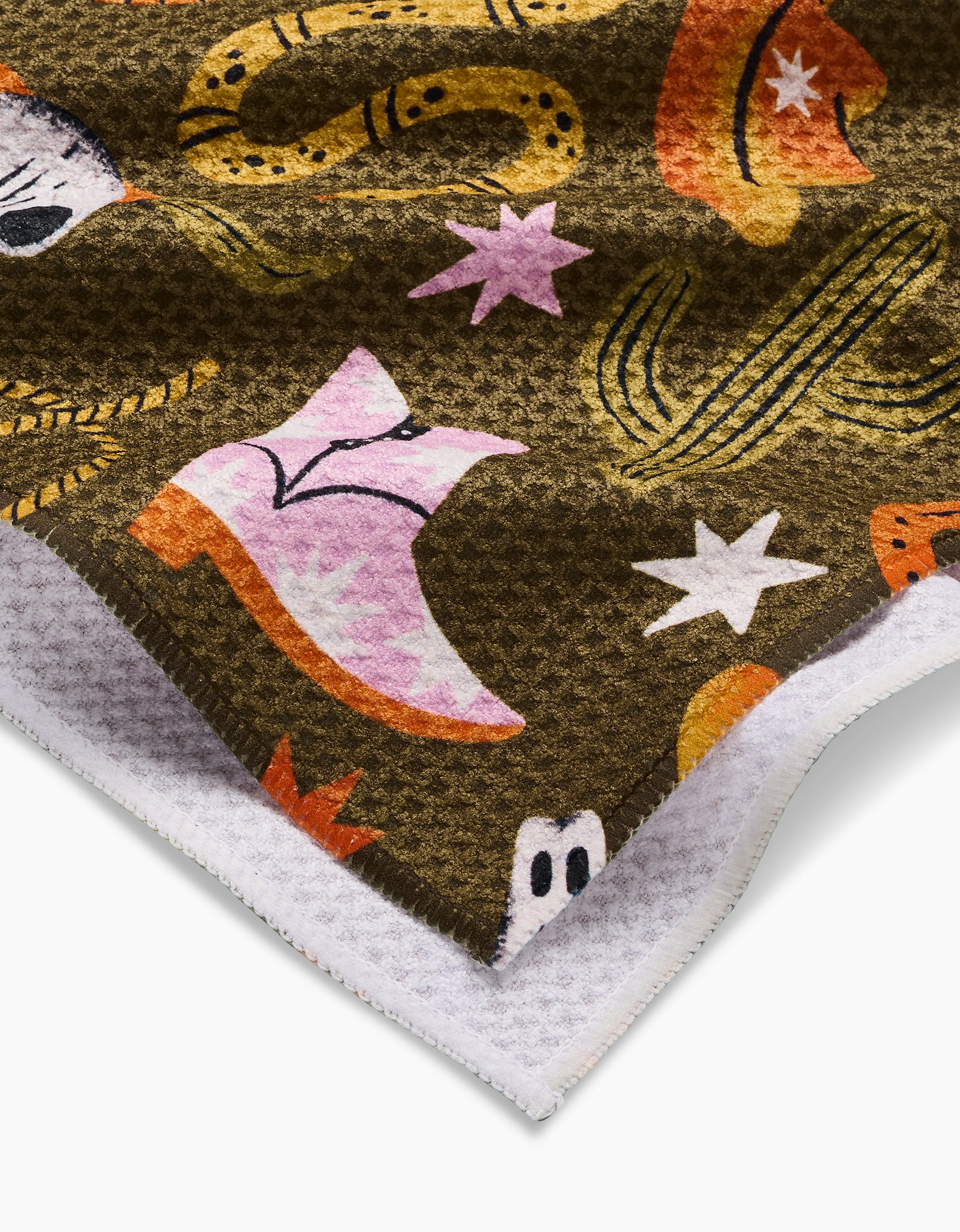 A close-up of the Megan Roy Wild Cowboy blanket, featuring colorful cowboy-themed designs—pink boots, orange hats, cacti, stars, and ghosts—on a dark brown Geoweave fabric with a soft white edge.