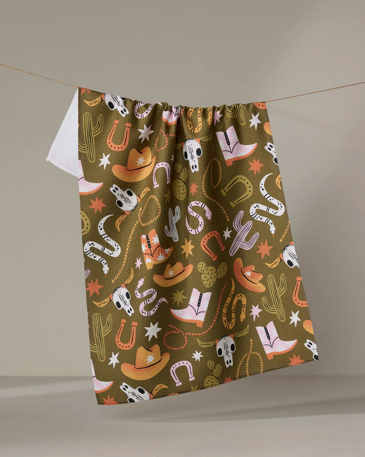 The "Wild Cowboy" tea towel by Megan Roy, made from recycled materials, features a colorful western pattern with cowboy hats, boots, cacti, horseshoes, snakes, stars, and bull skulls. It hangs on a line against a plain background.