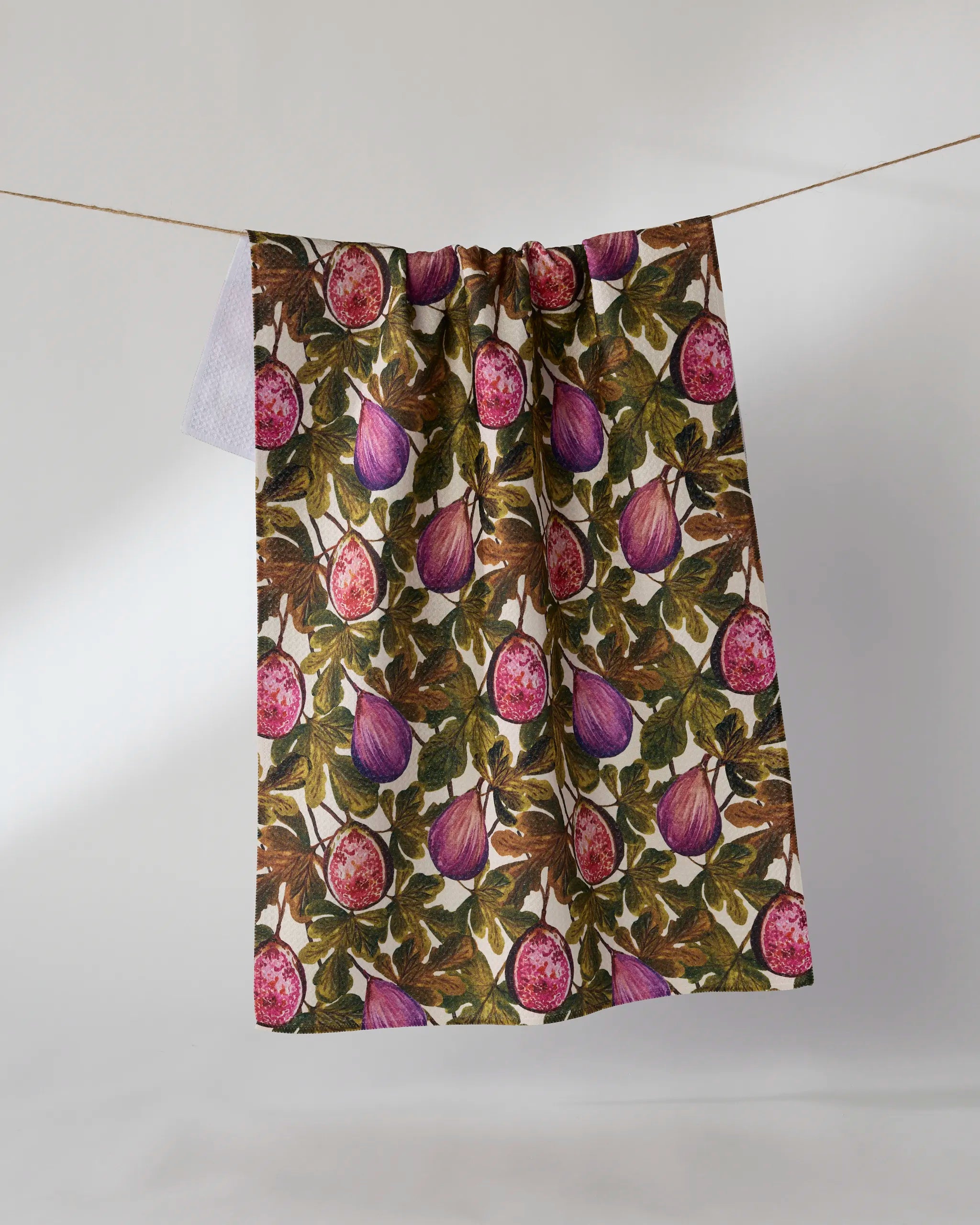 The Geometry Wild Fig tea towel, made from recycled materials and showcasing a vibrant fig and leaf design, hangs on a clothesline against a plain light gray background.