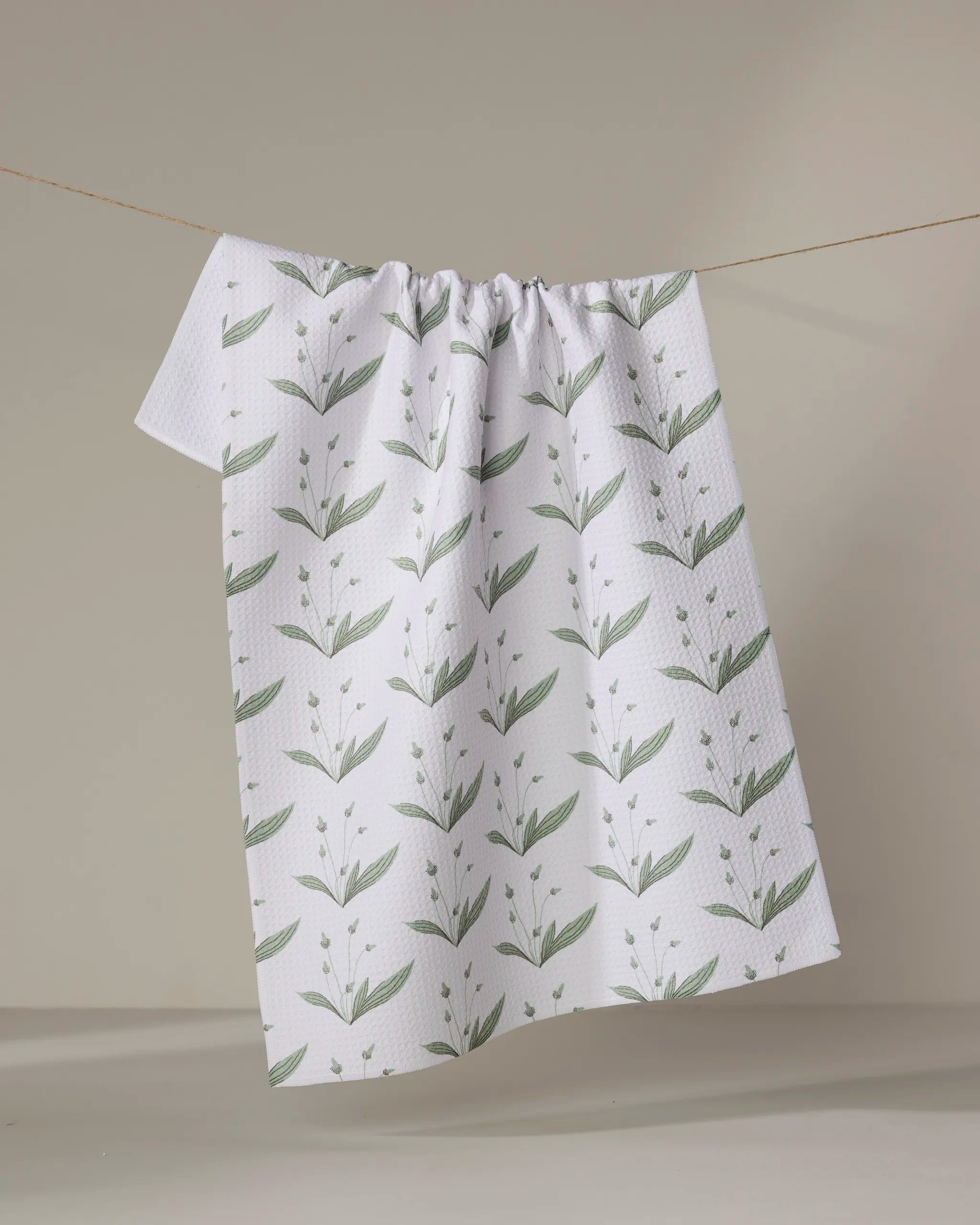 The Linda Fox Wild Leaves Green eco-friendly kitchen towel, featuring a white textured fabric and green botanical leaf pattern, hangs draped over a thin clothesline against a neutral background.
