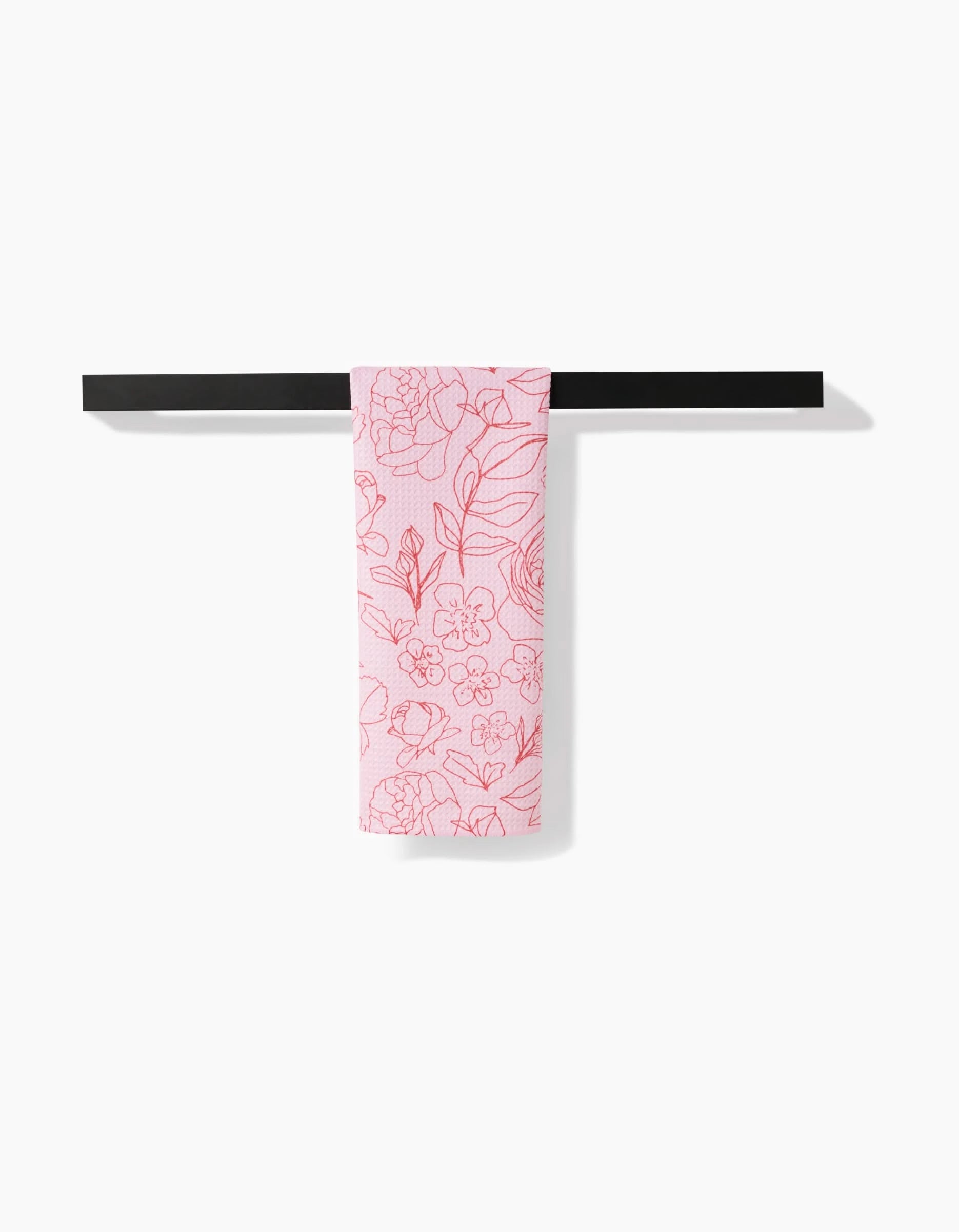 The Wild Roses tea towel by Janna Sue Design, featuring a pink floral pattern on white and made from recycled materials, is draped over a black towel bar against a plain white background.