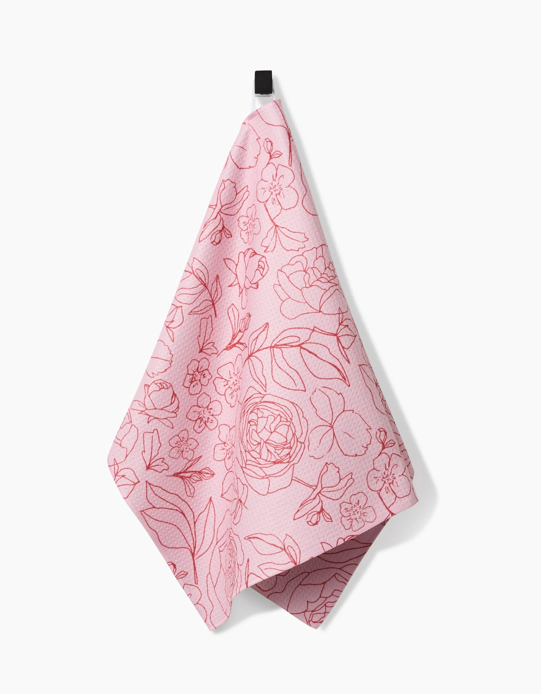 The Wild Roses kitchen towel by Janna Sue Design, made from recycled materials, features a pink background with a red floral line pattern and hangs from a black hook against a white backdrop.