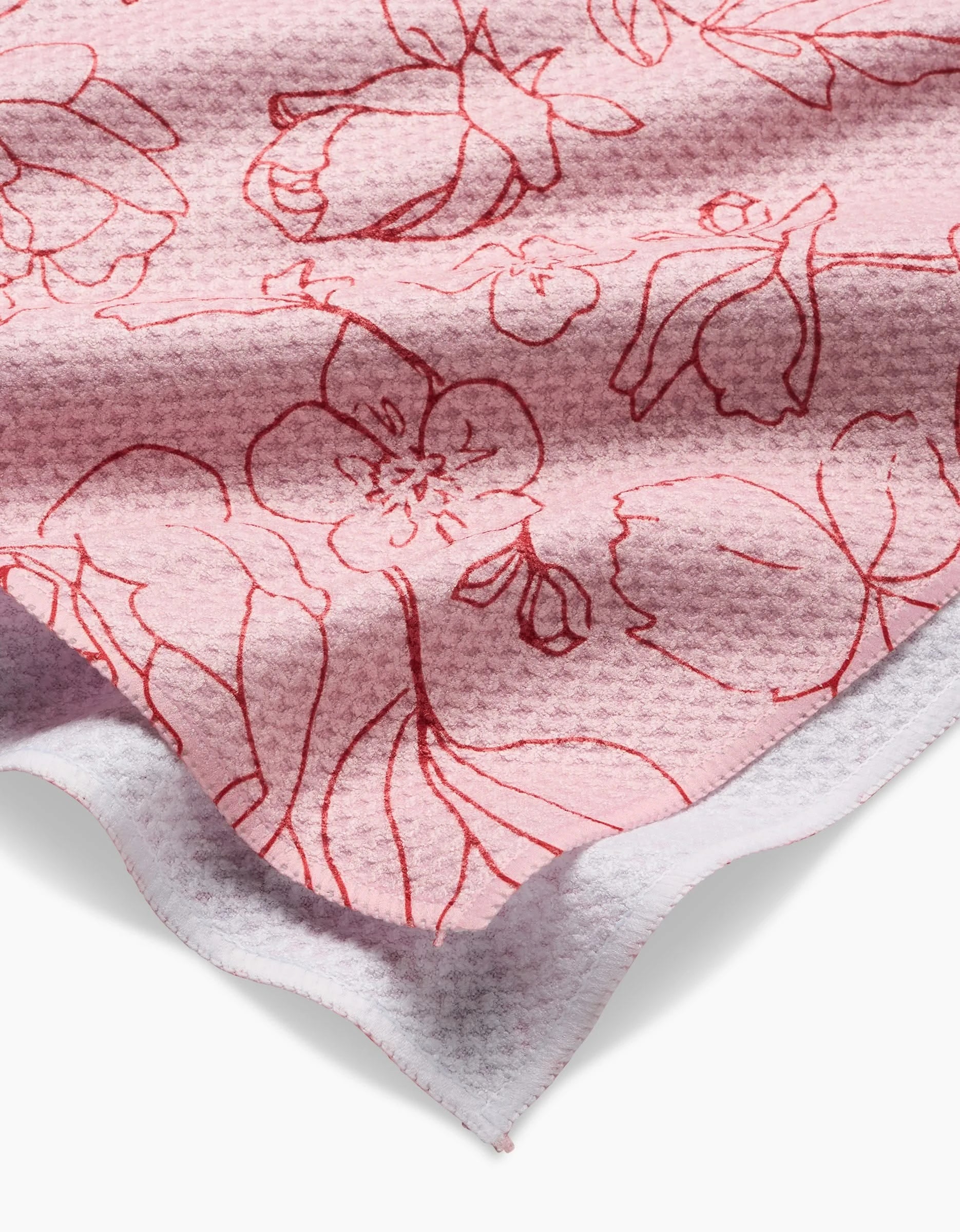 A close-up of Wild Roses by Janna Sue Design: a textured pink kitchen towel with a subtle waffle pattern, red floral line drawings, visible white underside, and wavy cut edge—crafted from recycled materials.