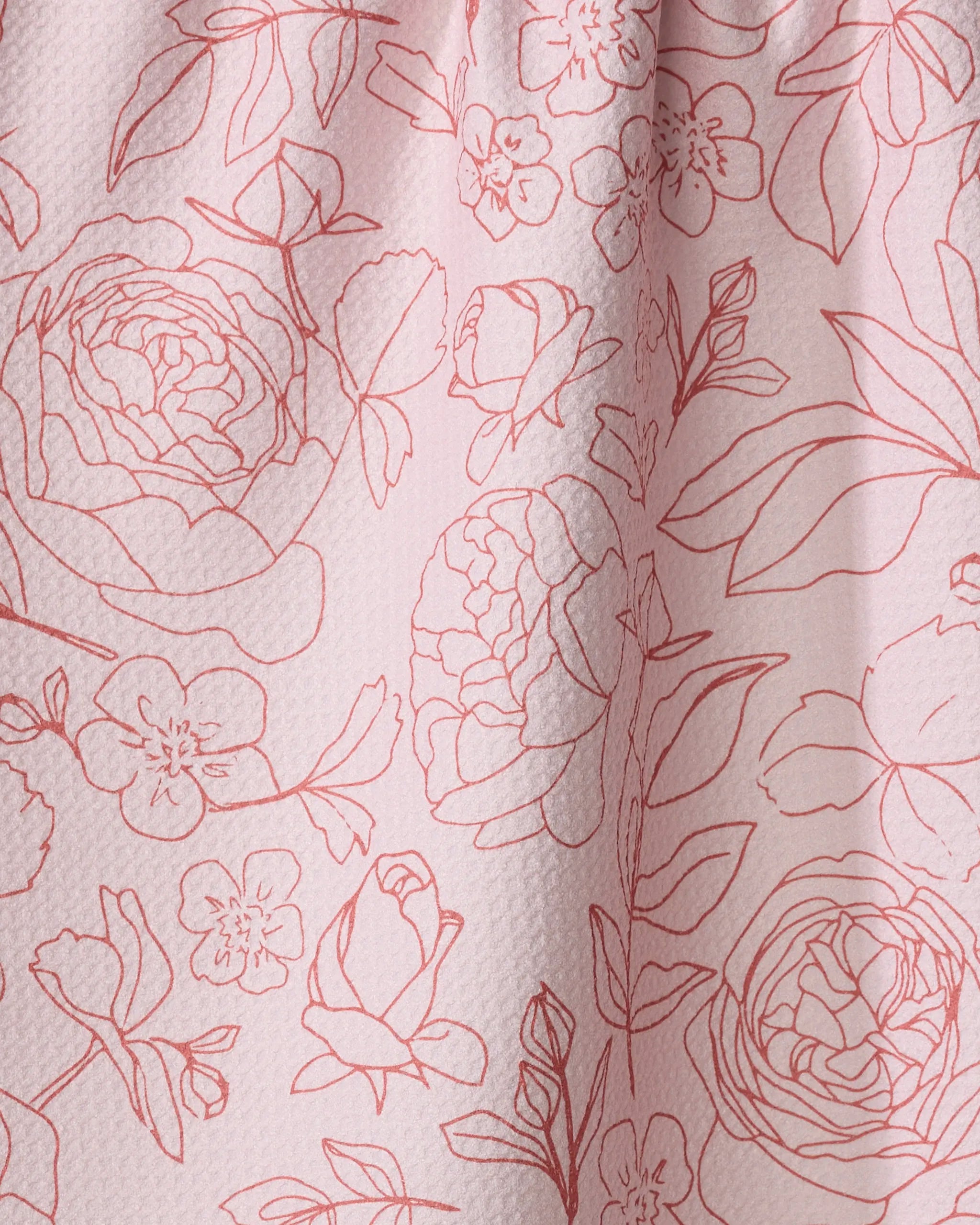 The "Wild Roses" dish towel by Janna Sue Design features pink textured fabric with a red floral outline of roses, peonies, leaves, and buds. Absorbent cotton and subtle folds highlight the elegant surface pattern.