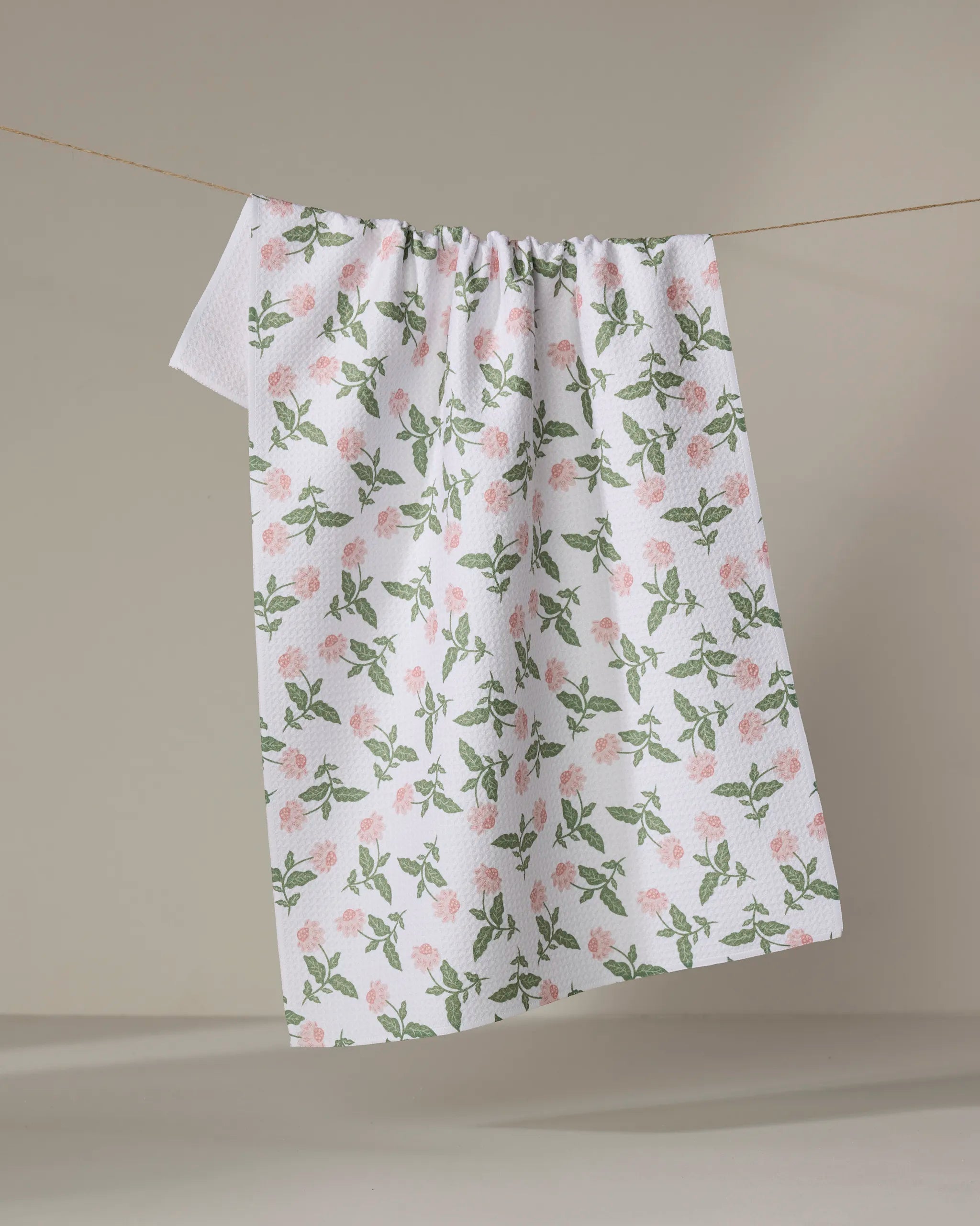 The Danelys Sidron "Wildflower Vine Blush" tea towel, featuring a pink rose and green leaf pattern, hangs on a clothesline against a plain beige background.