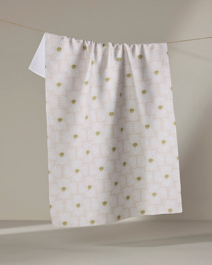 The Ash Cascade Wildflower tea towel, featuring a white textured design with subtle geometric patterns and green floral accents, hangs on a clothesline against a neutral backdrop.