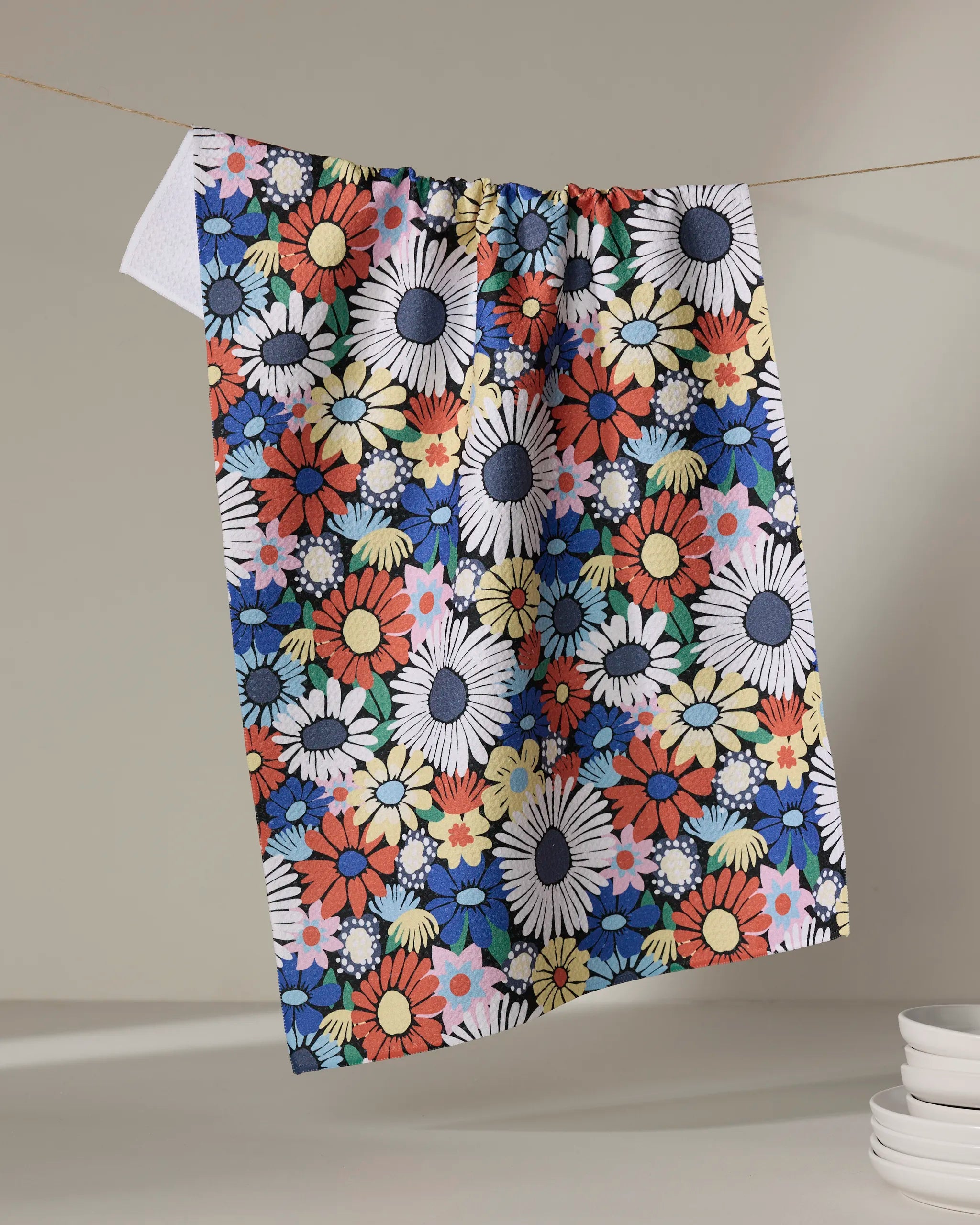 The Claire Iglesias Wildflowers Bouquet tea towel, featuring a vibrant floral pattern with large daisies in red, yellow, blue, white, and pink, hangs over a string against a neutral background with plates stacked nearby.
