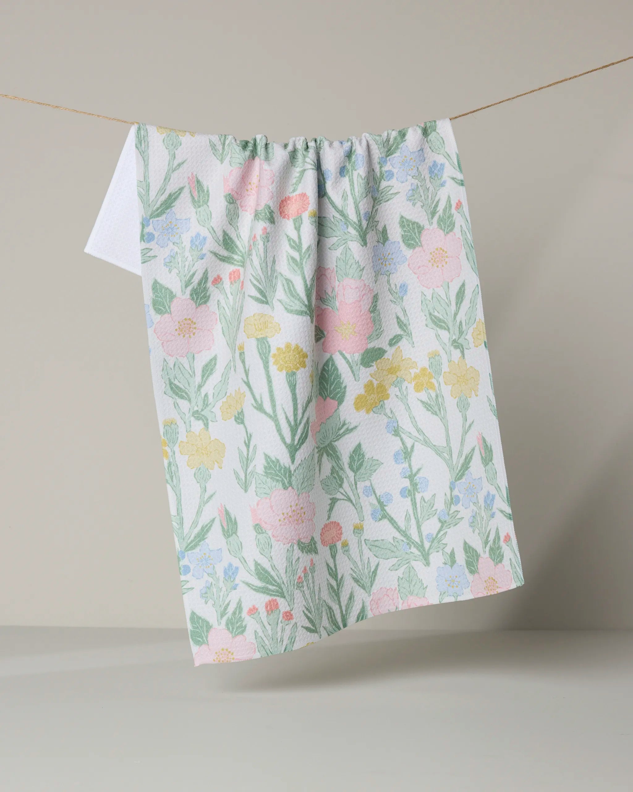 The Wildflowers in Bloom tea towel by Jasmine Blooms Designs hangs from a thin string against a light beige background, featuring pastel green leaves and pink, yellow, and blue floral patterns.