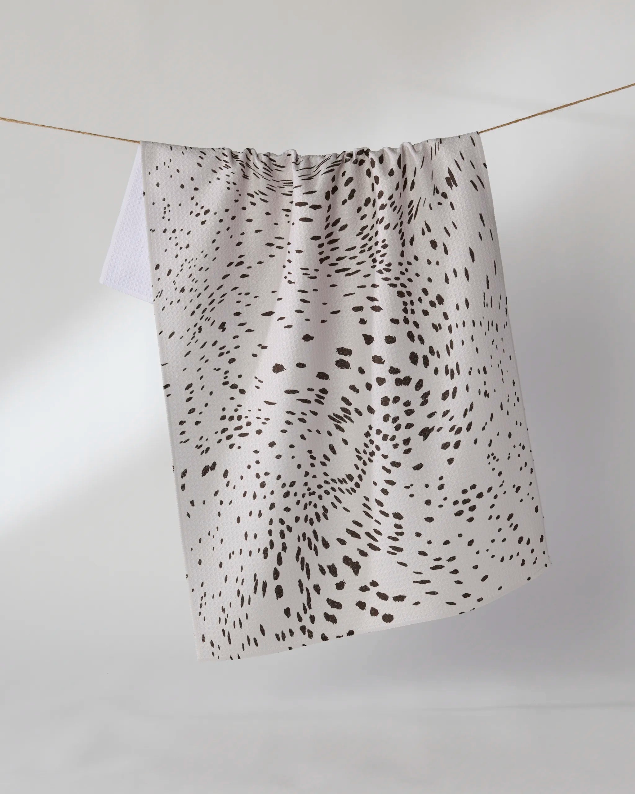 The Geometry Wildgrain white eco-friendly kitchen towel features an abstract scatter of black dots and hangs on a clothesline against a plain light background.