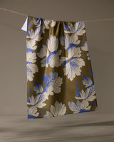 The Geometry Wildshade tea towel, featuring white and blue leaf patterns on a brown background, hangs from a string against a neutral backdrop. Its fabric dries quickly and adds style to any kitchen.