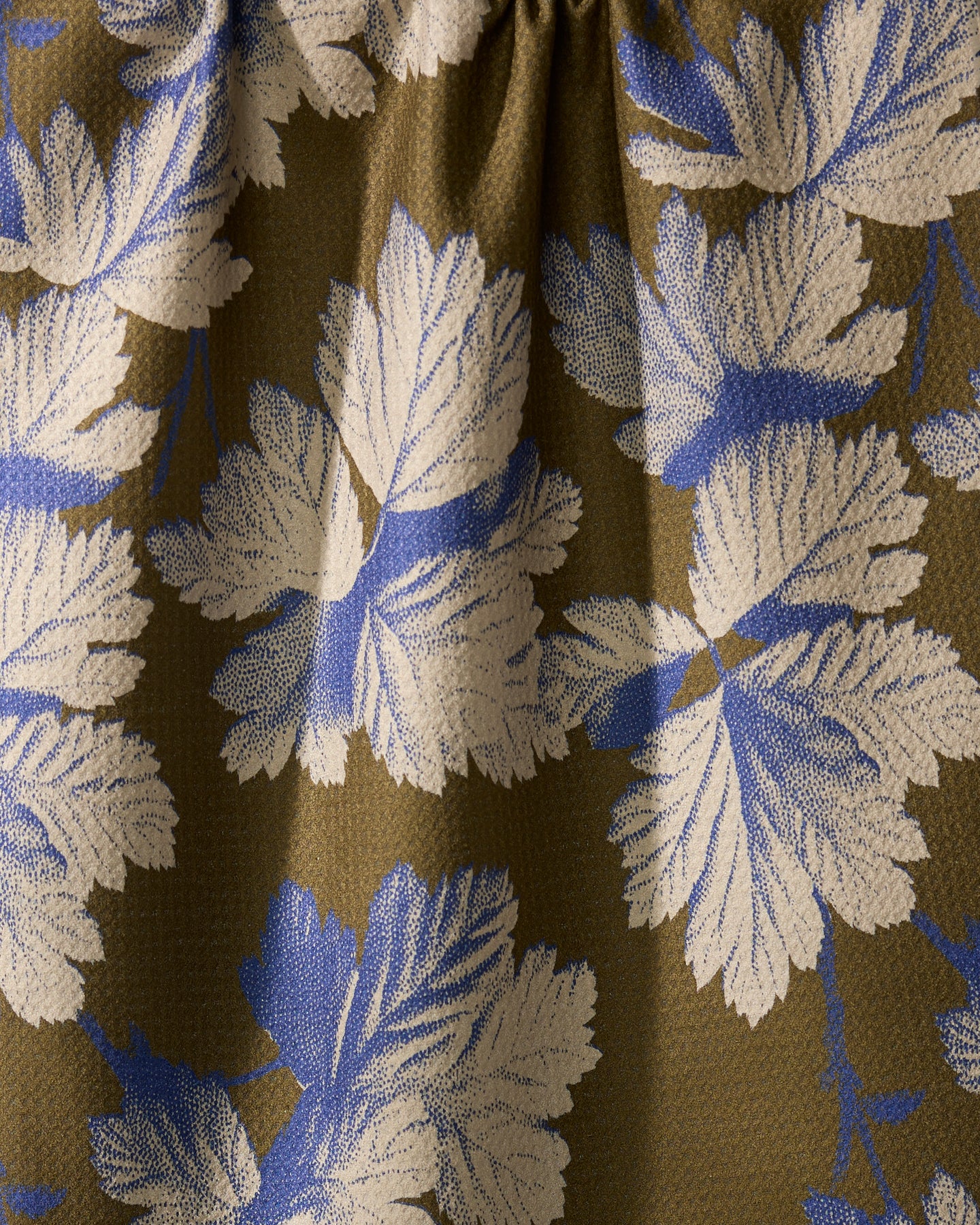 A close-up of Geometry’s Wildshade quick-drying fabric shows textured folds and shadows, displaying large white and blue leaves against an olive-green background.