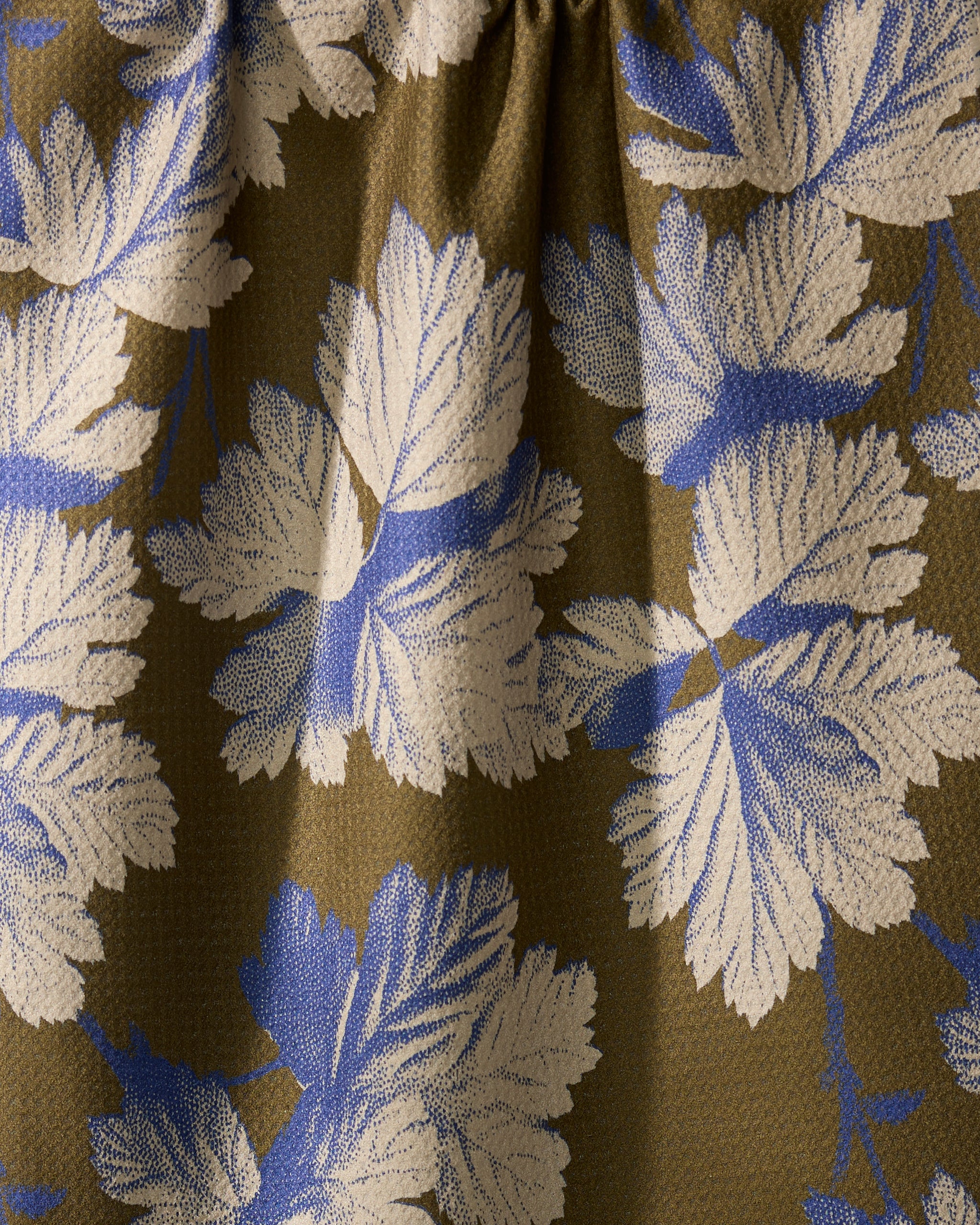 A close-up of Geometry’s Wildshade quick-drying fabric shows textured folds and shadows, displaying large white and blue leaves against an olive-green background.