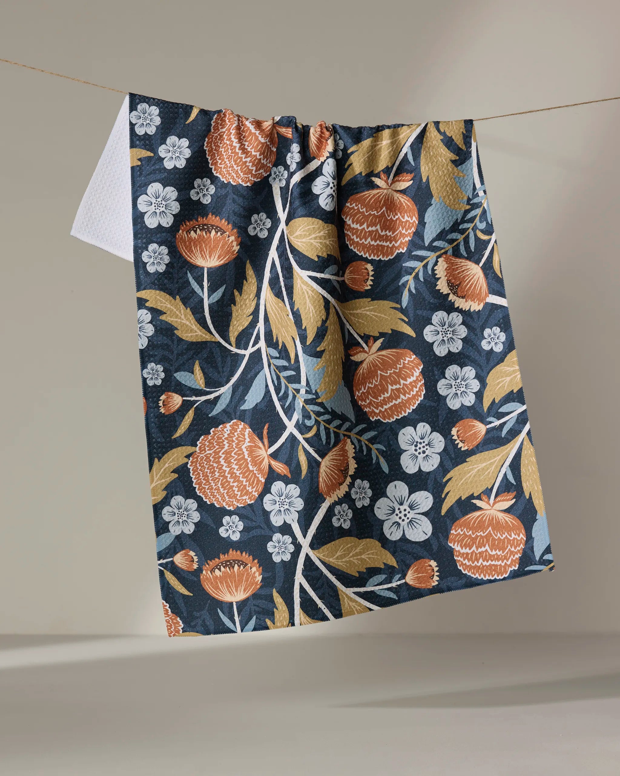 The William kitchen towel by Danelys Sidron features a bold orange, white, and mustard yellow floral pattern on dark blue, hanging on a string against a softly lit neutral background.