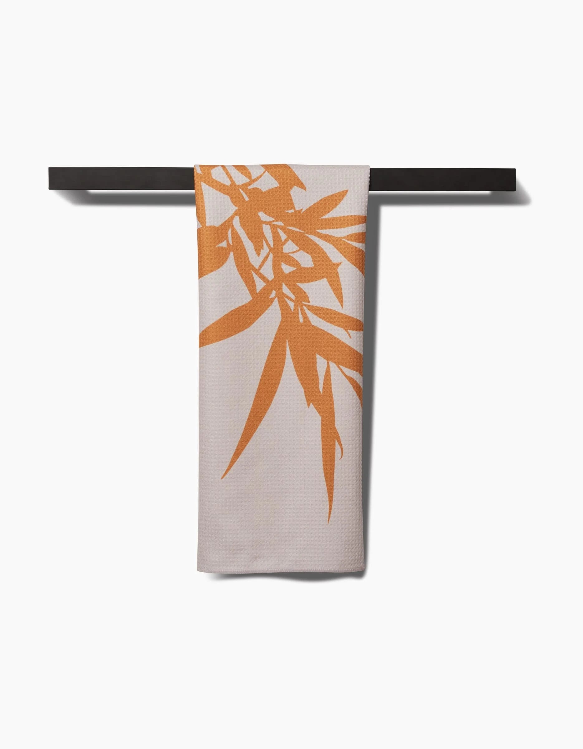 The PrintedPlant Willow tea towel, featuring an orange bamboo leaf design on beige Geoweave fabric, hangs neatly on a dark bar against a white background.