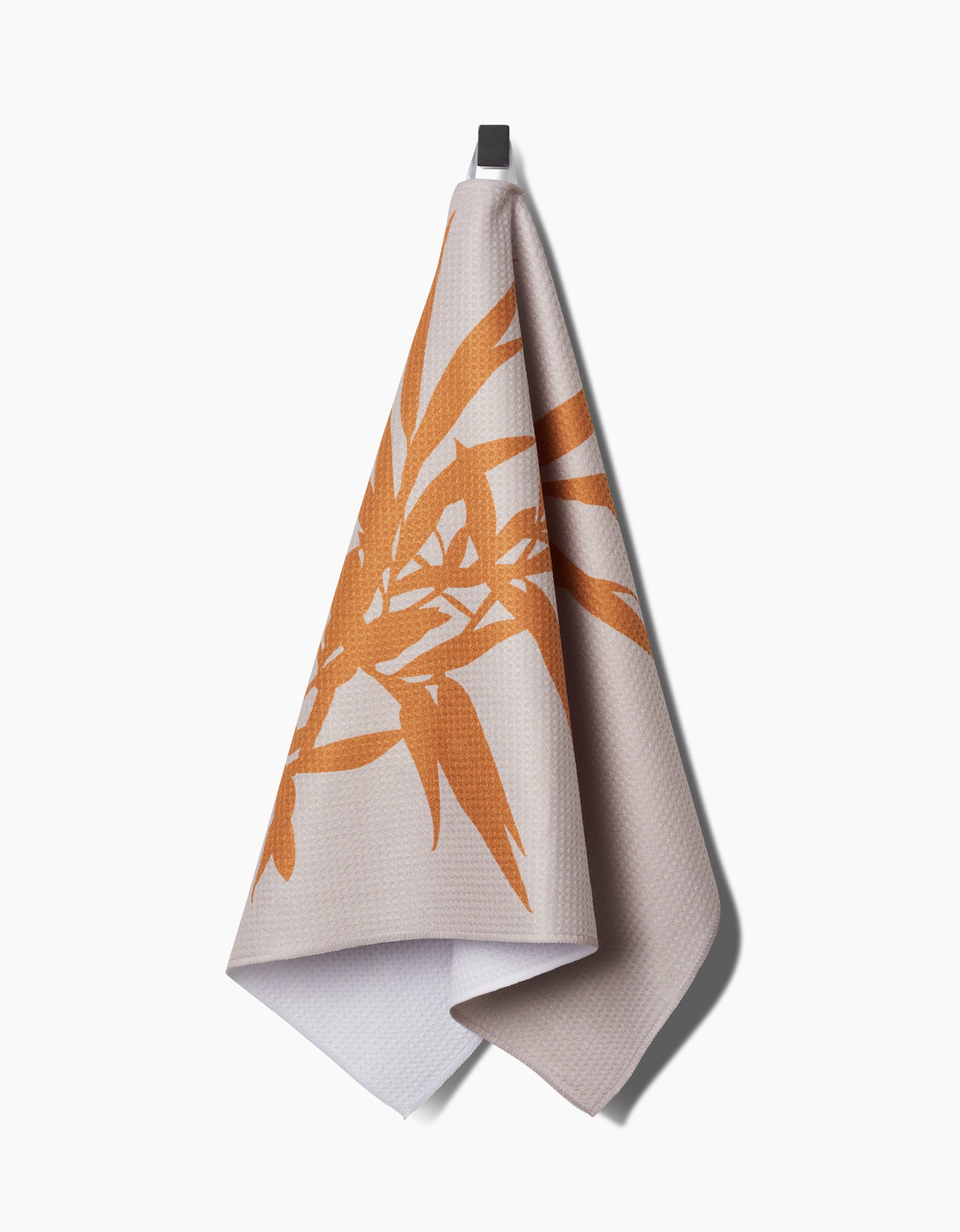 The PrintedPlant Willow tea towel, featuring an orange leaf design on beige and made from recycled materials, hangs from a black hook against a white background.