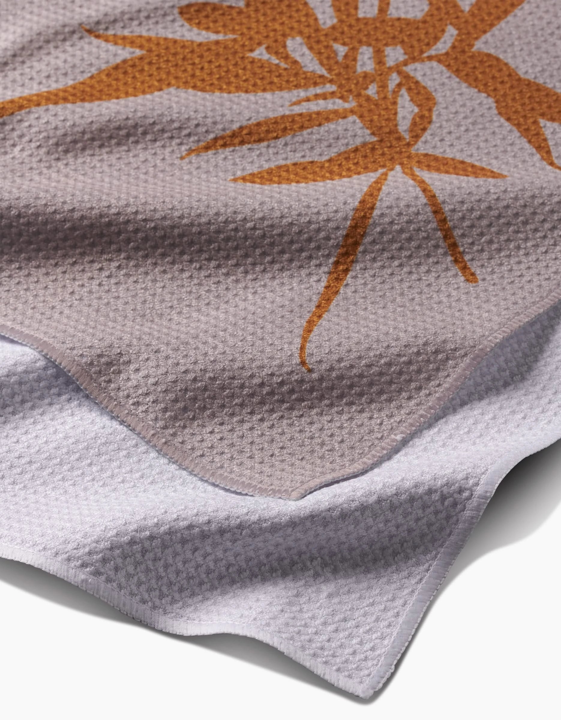 A close-up of two Willow tea towels by PrintedPlant: the top features a bold orange floral design on light brown, while the bottom is plain white, both with a textured waffle-weave.
